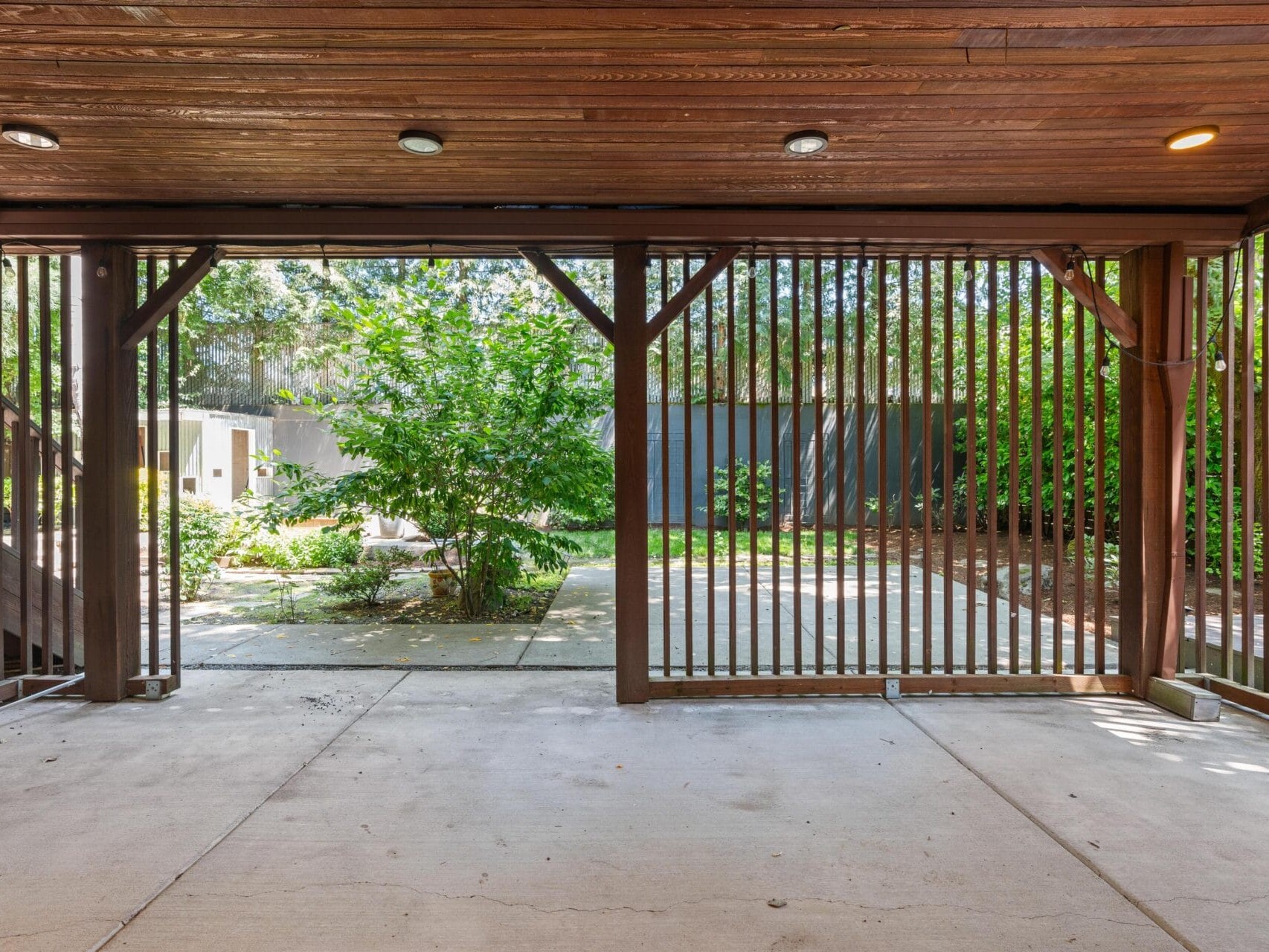 Covered patio with a wooden ceiling and vertical wooden slats, overlooking a backyard with green trees, a concrete path, and a bench. Natural light filters through the open space.
