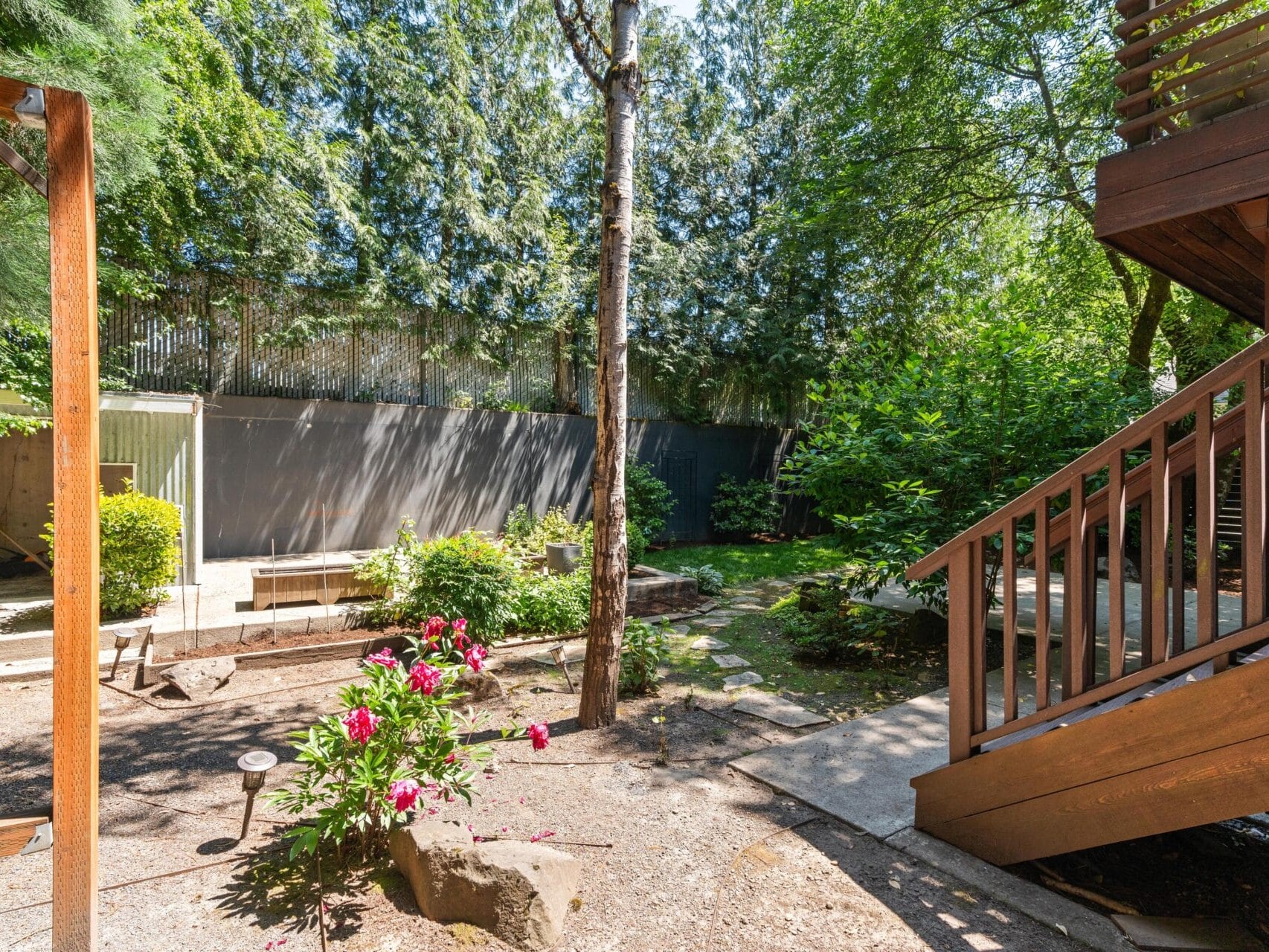 A sunlit backyard features blooming flowers, trees, green shrubs, a wooden staircase, and a privacy fence. Shadows and dappled sunlight fall across the garden and pathway.