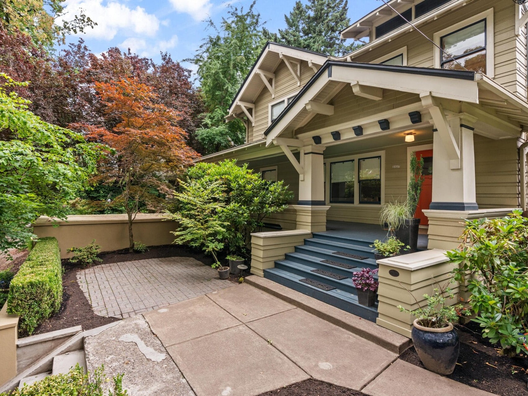 A beige Craftsman-style house with a covered front porch, blue steps, and red front door, surrounded by lush green trees and landscaped plants, with a paved walkway and driveway.