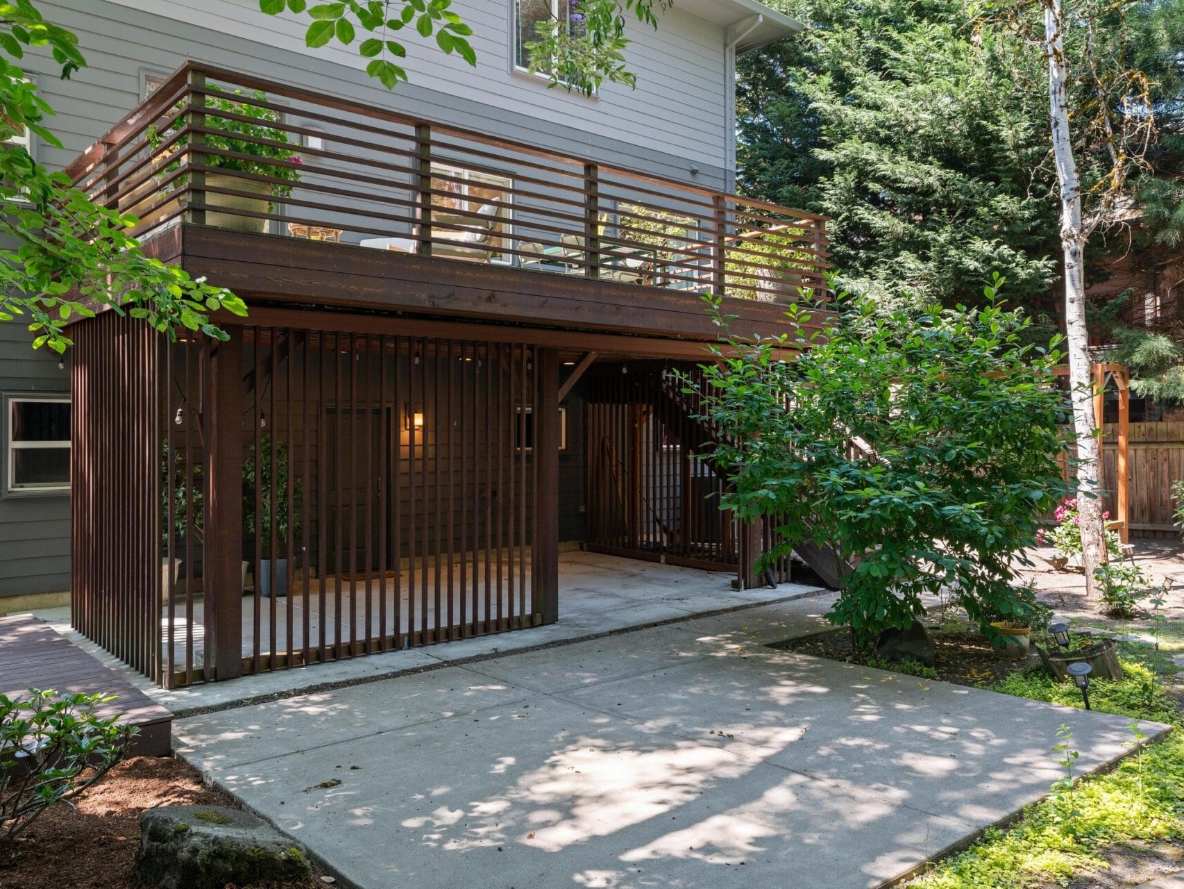A modern two-story house with a wooden deck and slatted privacy panels overlooks a shaded patio surrounded by green trees and plants.