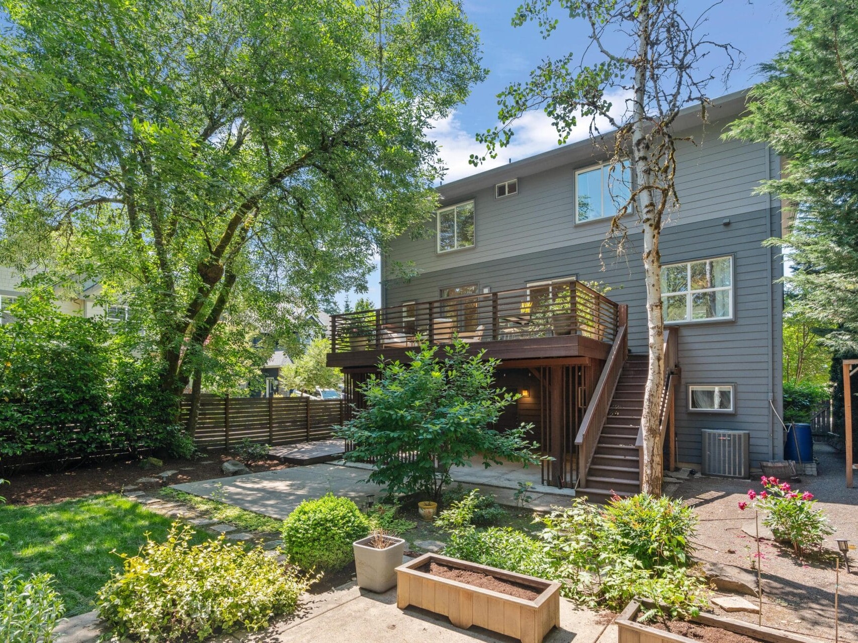 A modern two-story gray house with a wooden balcony and outdoor staircase, surrounded by trees, green lawn, potted plants, and a landscaped backyard on a sunny day.