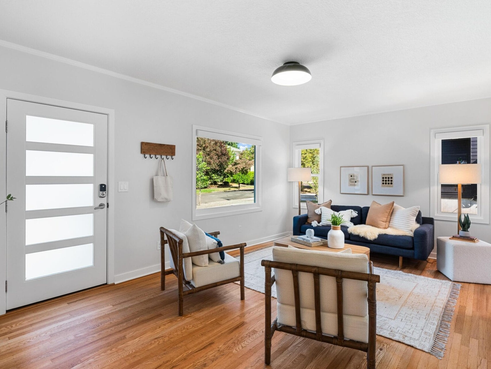 Bright, modern living room with wooden floors, white walls, large windows, a navy sofa, two wooden armchairs, a coffee table, and wall art. Natural light fills the space, creating a cozy and inviting atmosphere.