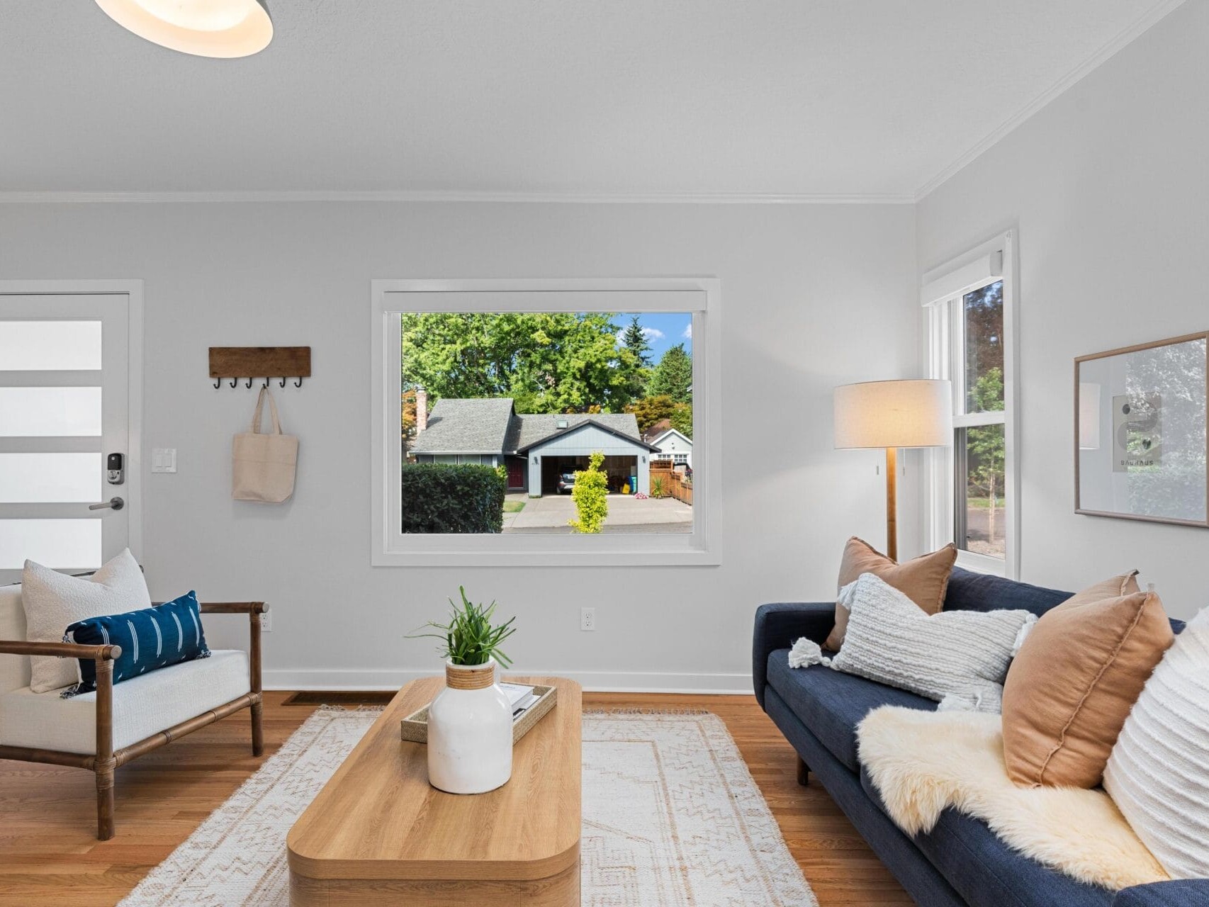 Bright living room with a modern sofa, armchair, and coffee table. Large window shows a view of a house and greenery outside. Light walls, wood floors, and cozy decor create a welcoming atmosphere.