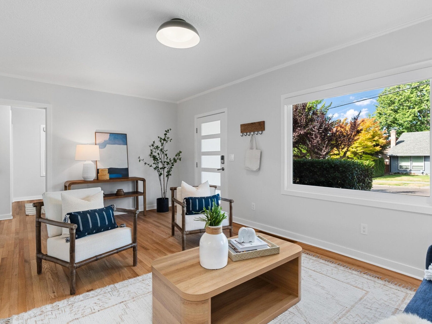 A bright, modern living room with white walls, wood flooring, two armchairs, a wooden coffee table, blue accents, and a large window showing a green, tree-filled yard outside.