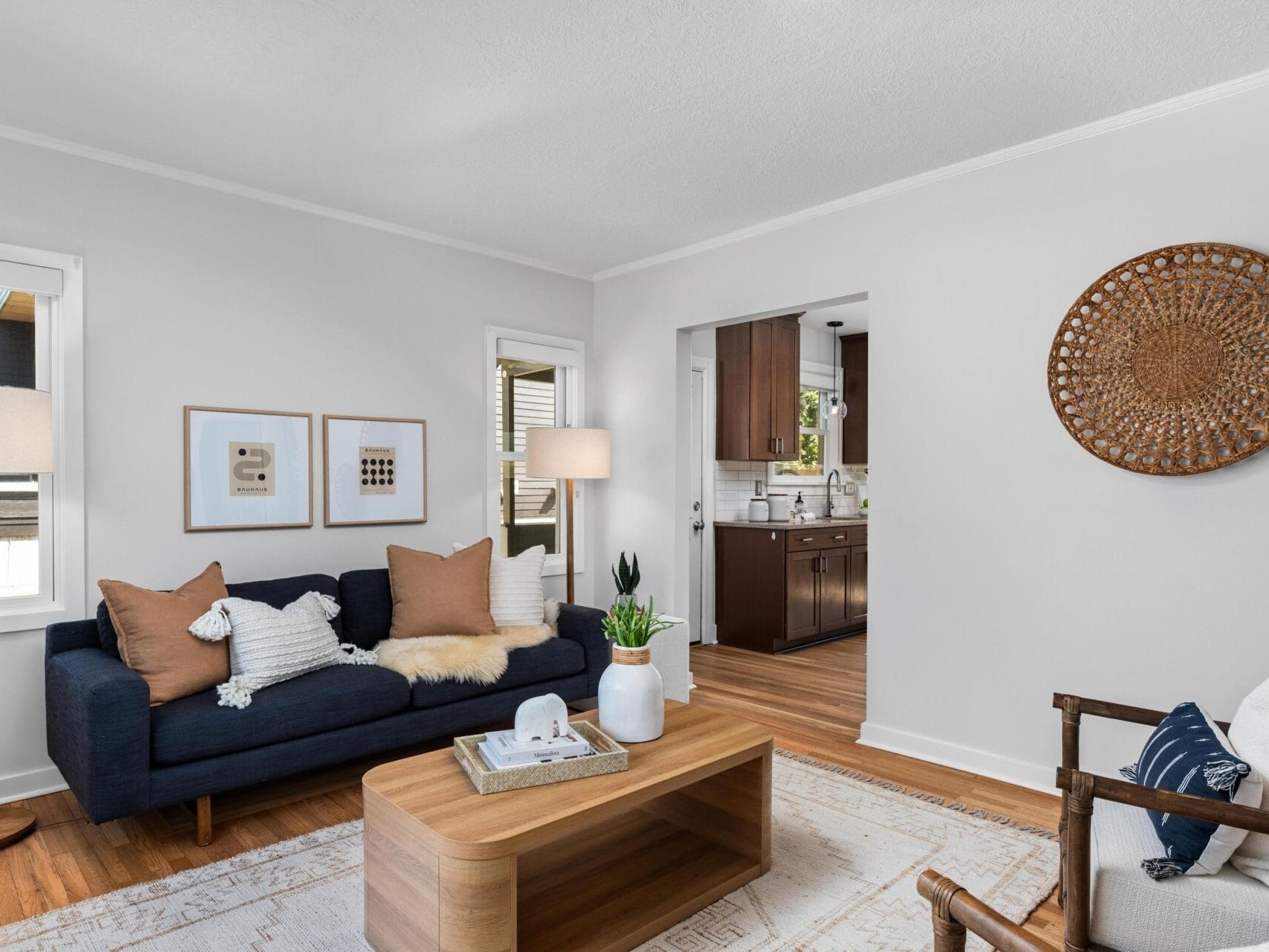 Modern living room with a dark blue sofa, neutral pillows, a wooden coffee table, a patterned rug, and a woven wall decor. A bright kitchen with wooden cabinets is visible through the doorway.