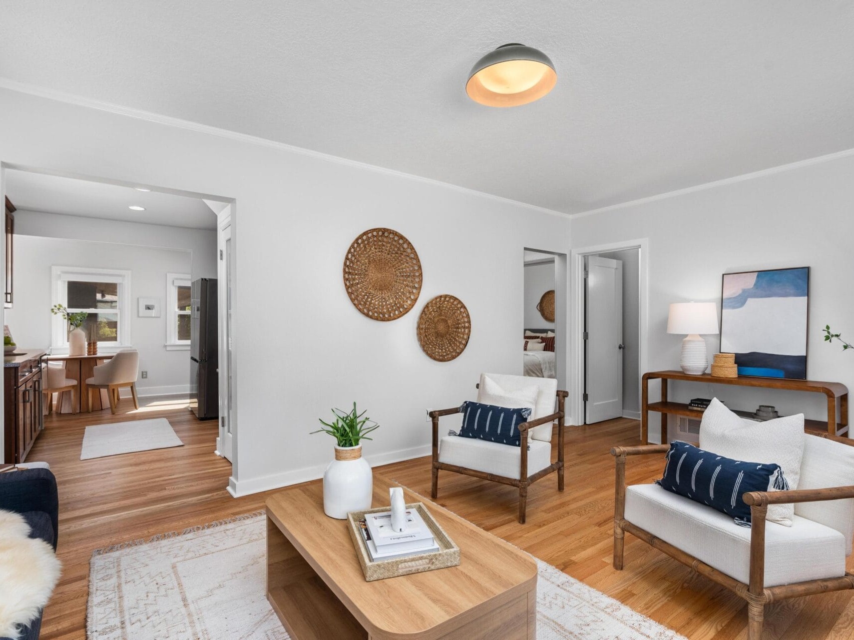 Bright living room with white walls, wood floors, a beige rug, wooden coffee table, two armchairs, a sofa, wall art, woven wall decor, and a sideboard with a lamp and plant. Kitchen is visible in the background.