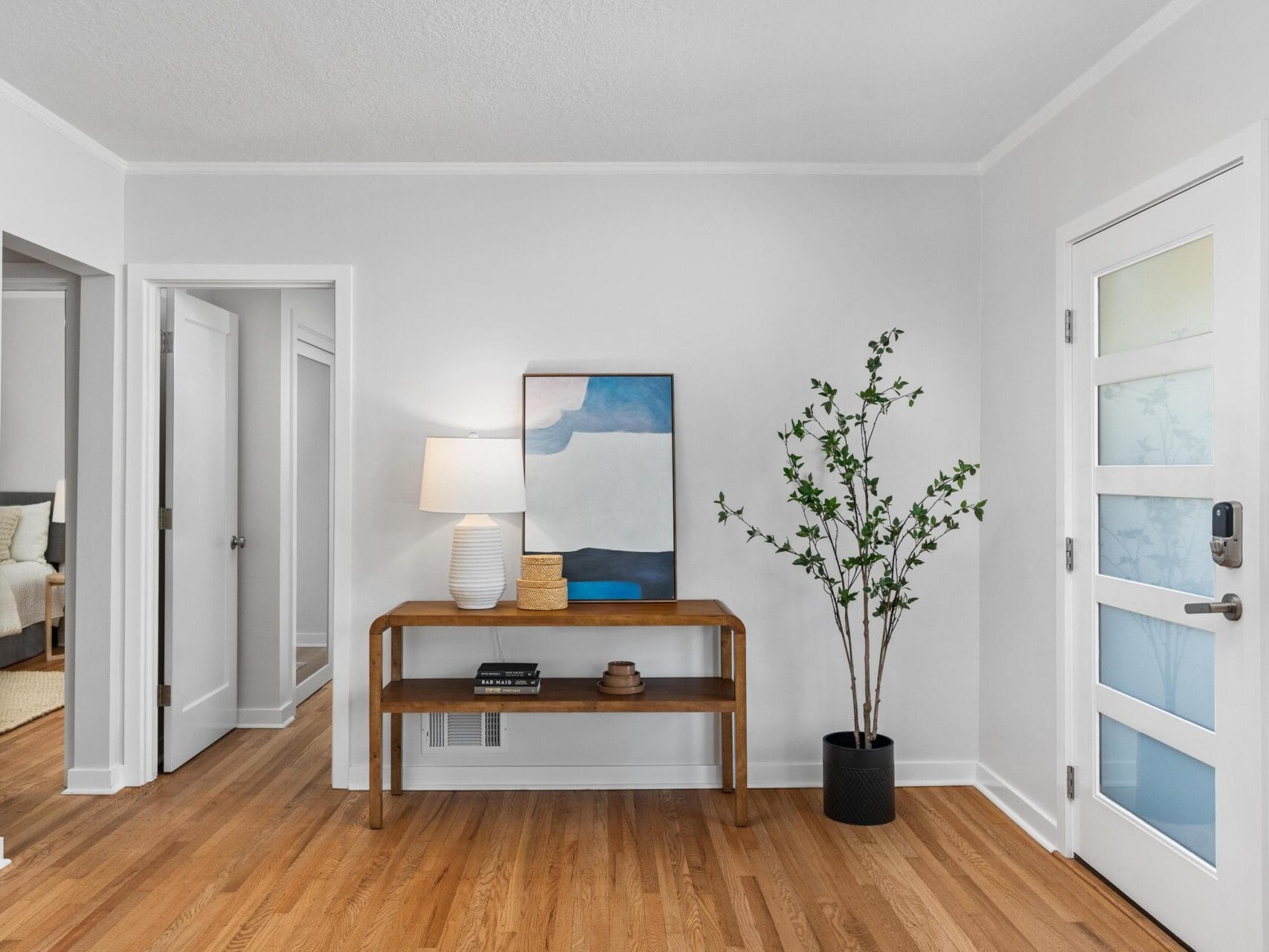 A modern, minimalist entryway with light wood floors, a wooden console table holding a lamp, books, and art, and a potted plant beside it. White walls and door with frosted glass panels complete the space.