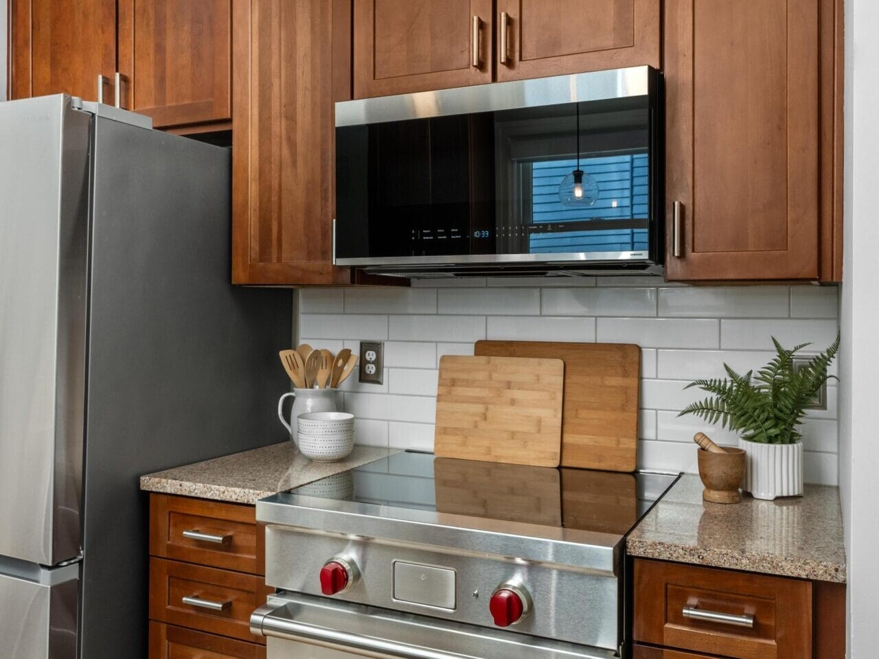 A modern kitchen features wooden cabinets, a stainless steel oven with red knobs, a microwave, a refrigerator, a potted plant, utensils in a holder, and two wooden cutting boards on a speckled countertop.