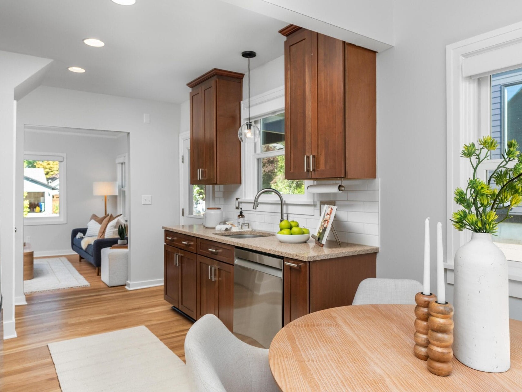 Modern kitchen with wooden cabinets, stainless steel appliances, and a round dining table with a vase and candles. Bright, natural light fills the space, with a cozy living area visible in the background.