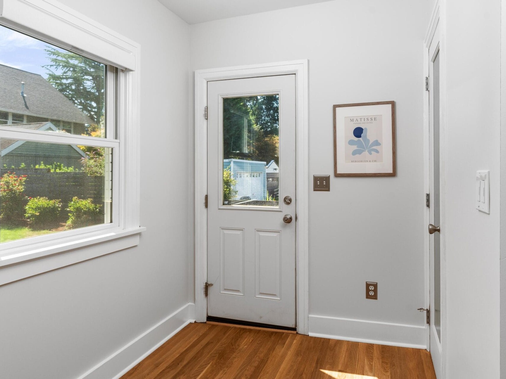 A bright, small entryway with white walls, a door with a window, a window to the left showing a green yard, a wooden floor, and a framed Matisse print on the wall.