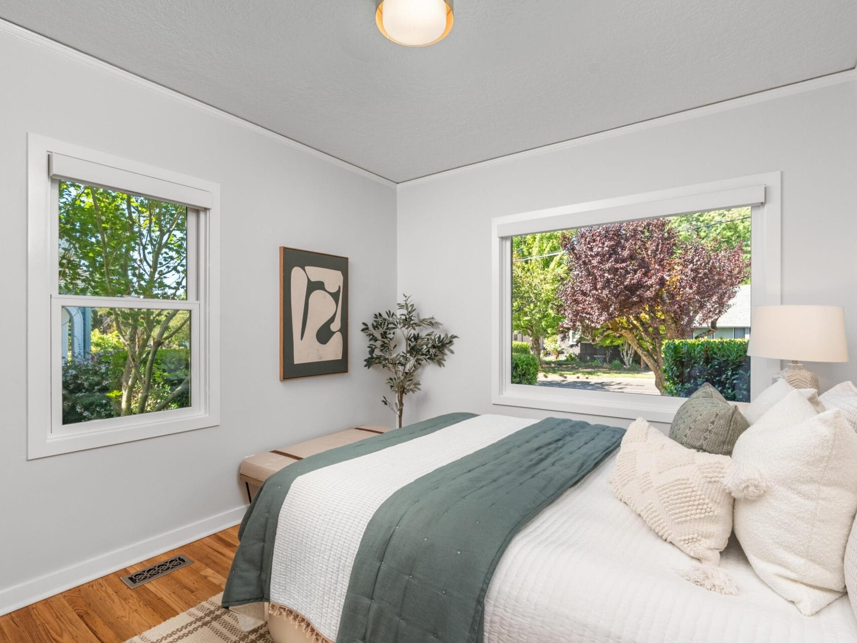 A bright bedroom with white walls, large windows showing green trees outside, a bed with white and green bedding, pillows, a light wooden bench, and a framed abstract artwork on the wall.