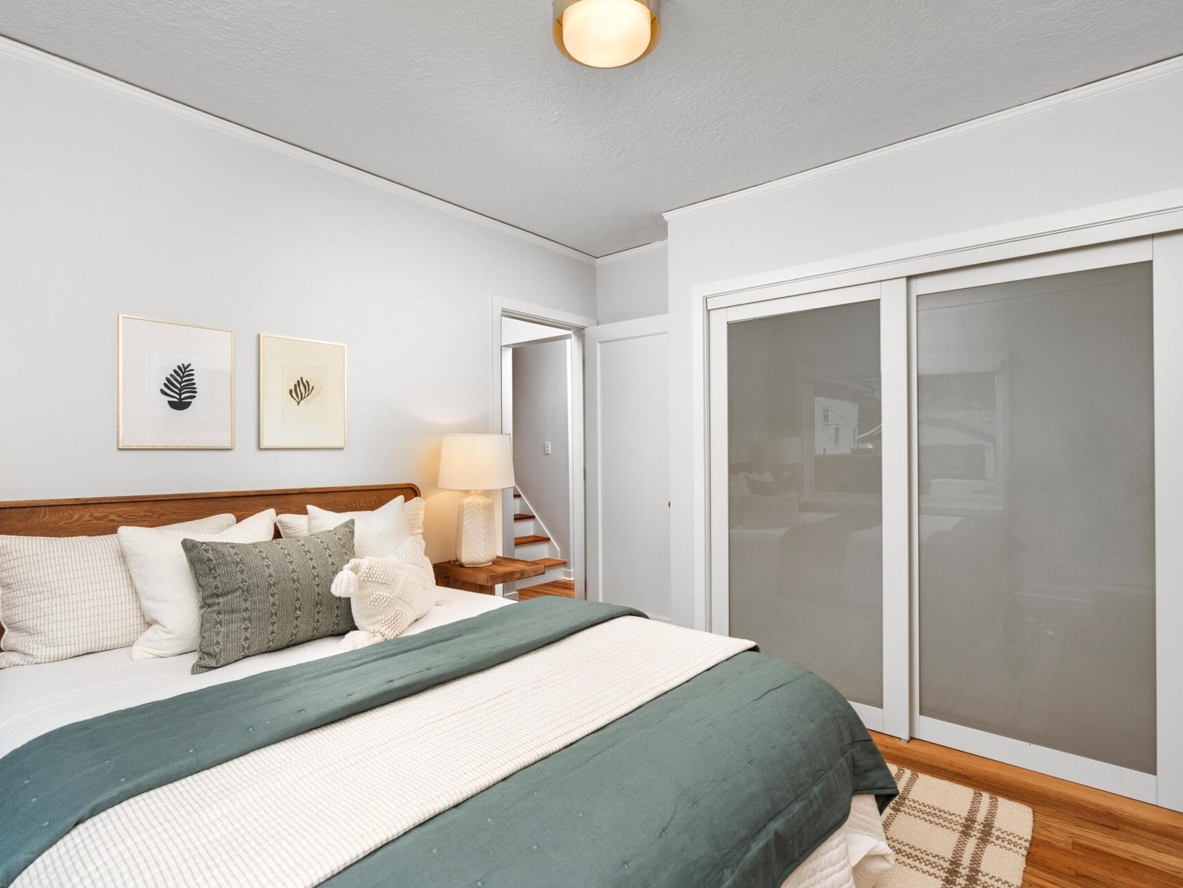 A modern bedroom with a wooden bed, white and green bedding, two framed prints on the wall, a plaid rug, sliding closet doors with frosted glass, and a small stairway visible through an open doorway.