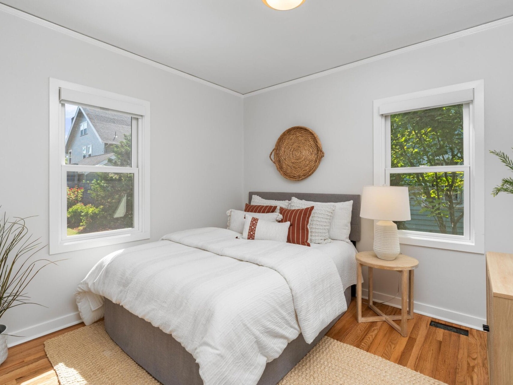 A cozy bedroom with a neatly made bed, white bedding, and striped pillows. There are two windows, a woven wall decor above the bed, a lamp on a wooden nightstand, and plants adding greenery to the space.