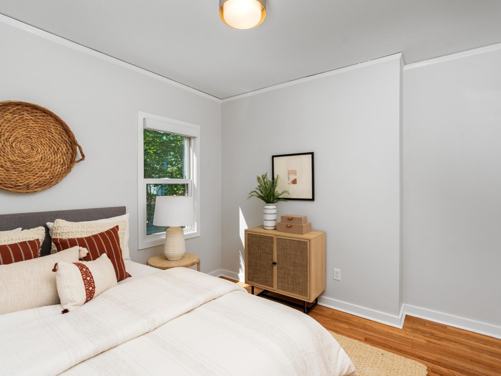 Bright bedroom with white walls, a bed with cream and rust pillows, a woven basket wall decor, a window, a small cabinet with plants and artwork, wood floor, and a neutral rug.
