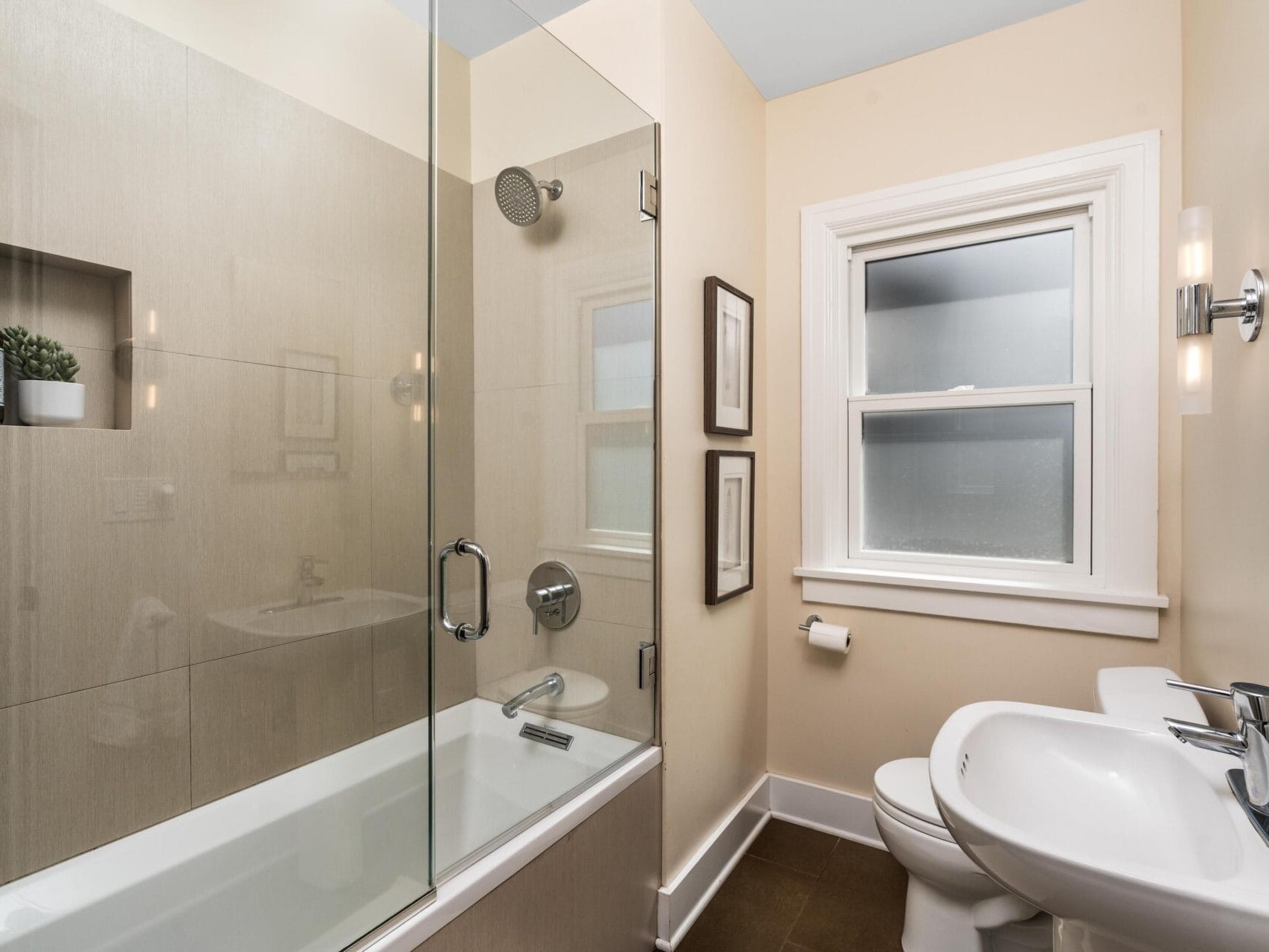 Modern bathroom with beige walls, a glass-enclosed bathtub and shower, white sink and toilet, frosted window, wall-mounted lights, and small shelves with decorative items.
