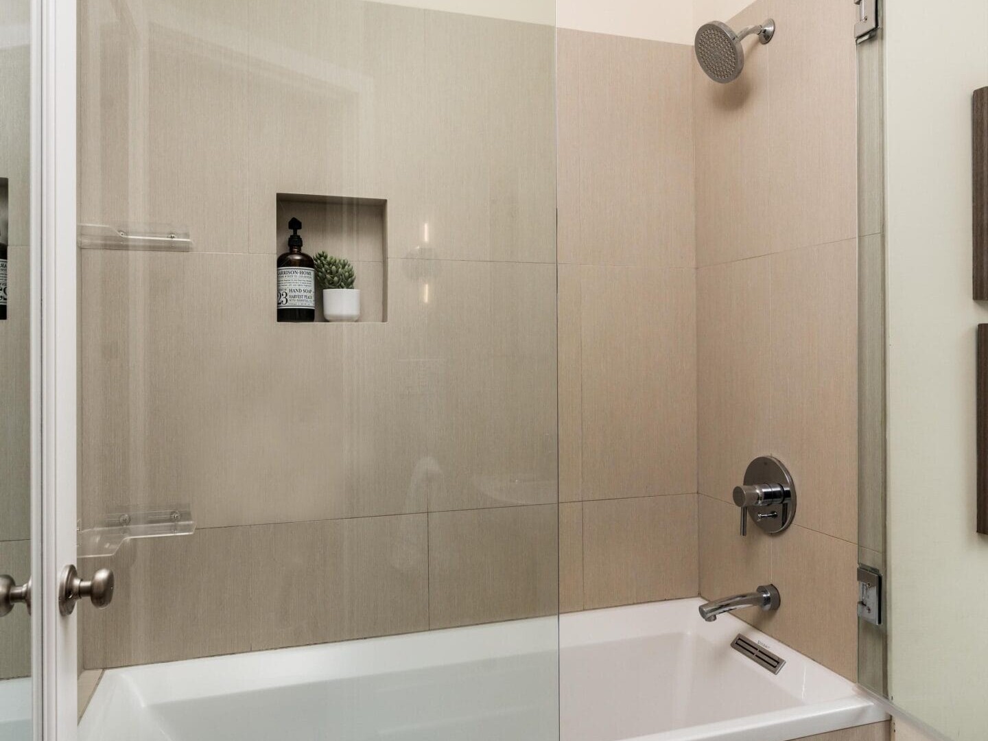 A modern bathroom with a glass shower screen over a white bathtub, beige tile walls, a wall-mounted showerhead, and a built-in niche holding a plant and toiletries.