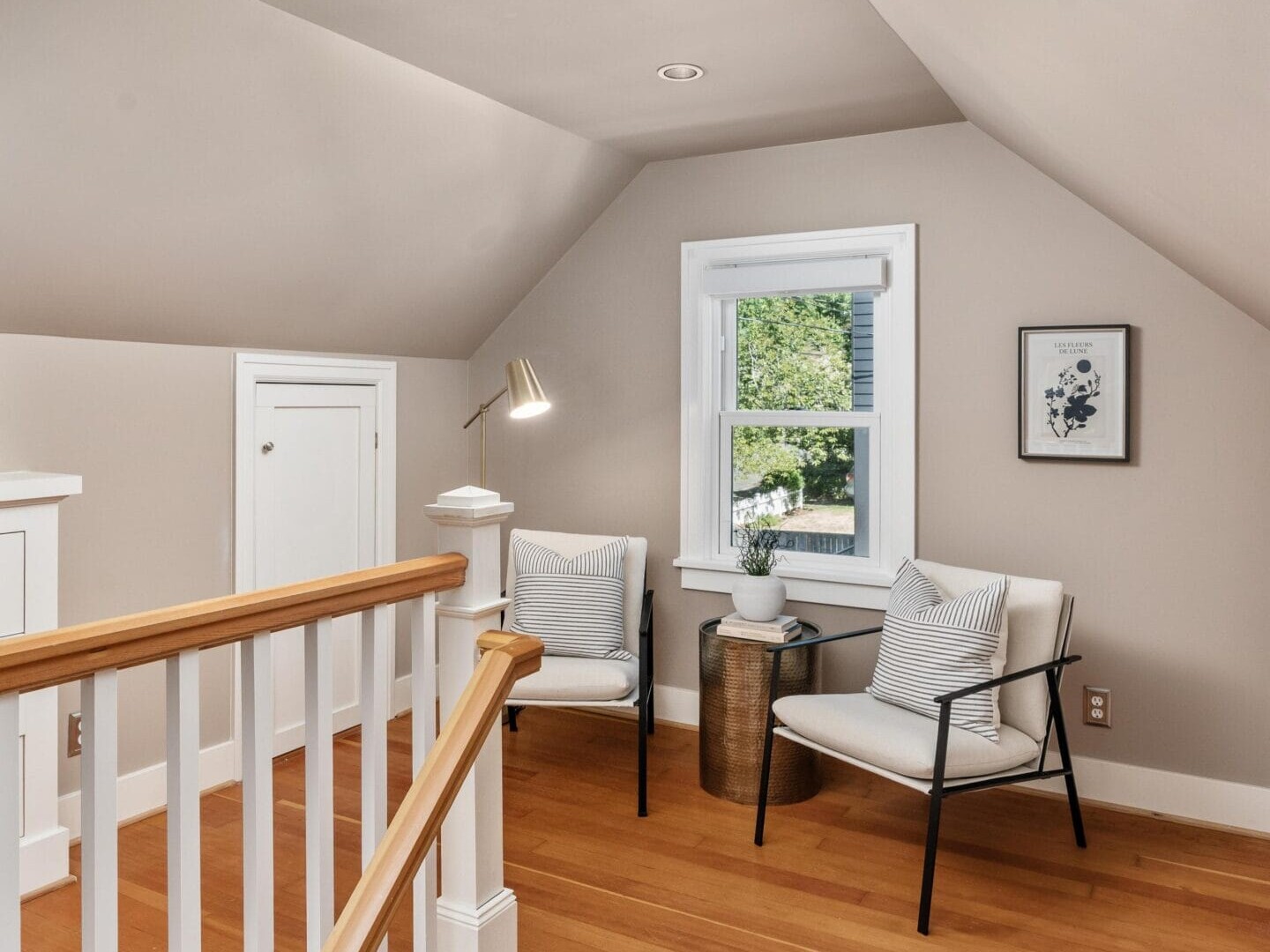 Small loft seating area with two modern chairs, a side table, a floor lamp, light-colored walls, hardwood floors, and a window letting in natural light, next to a staircase with white railing.
