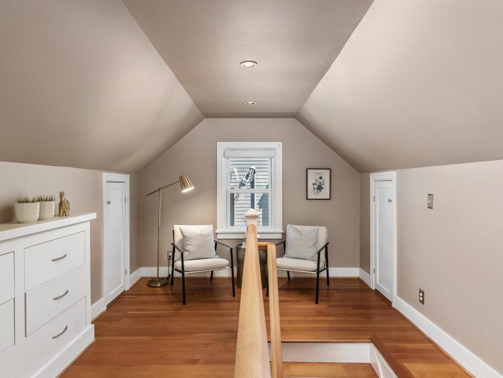 A cozy attic nook with sloped ceilings, two armchairs and a small table by a window, a floor lamp, white built-in drawers, wood flooring, and minimalist decor.