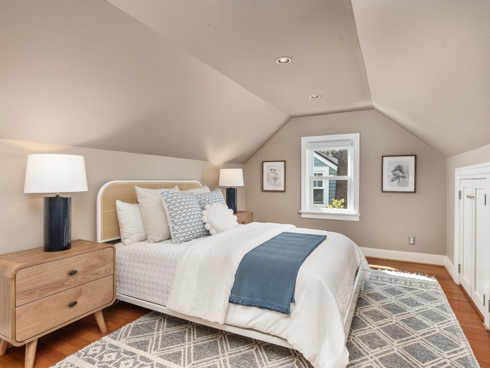 Cozy attic bedroom with sloped ceiling, a neatly made bed with white and blue bedding, two wooden nightstands with lamps, framed art above the bed, and built-in cabinets on the right wall. Large rug covers hardwood floor.