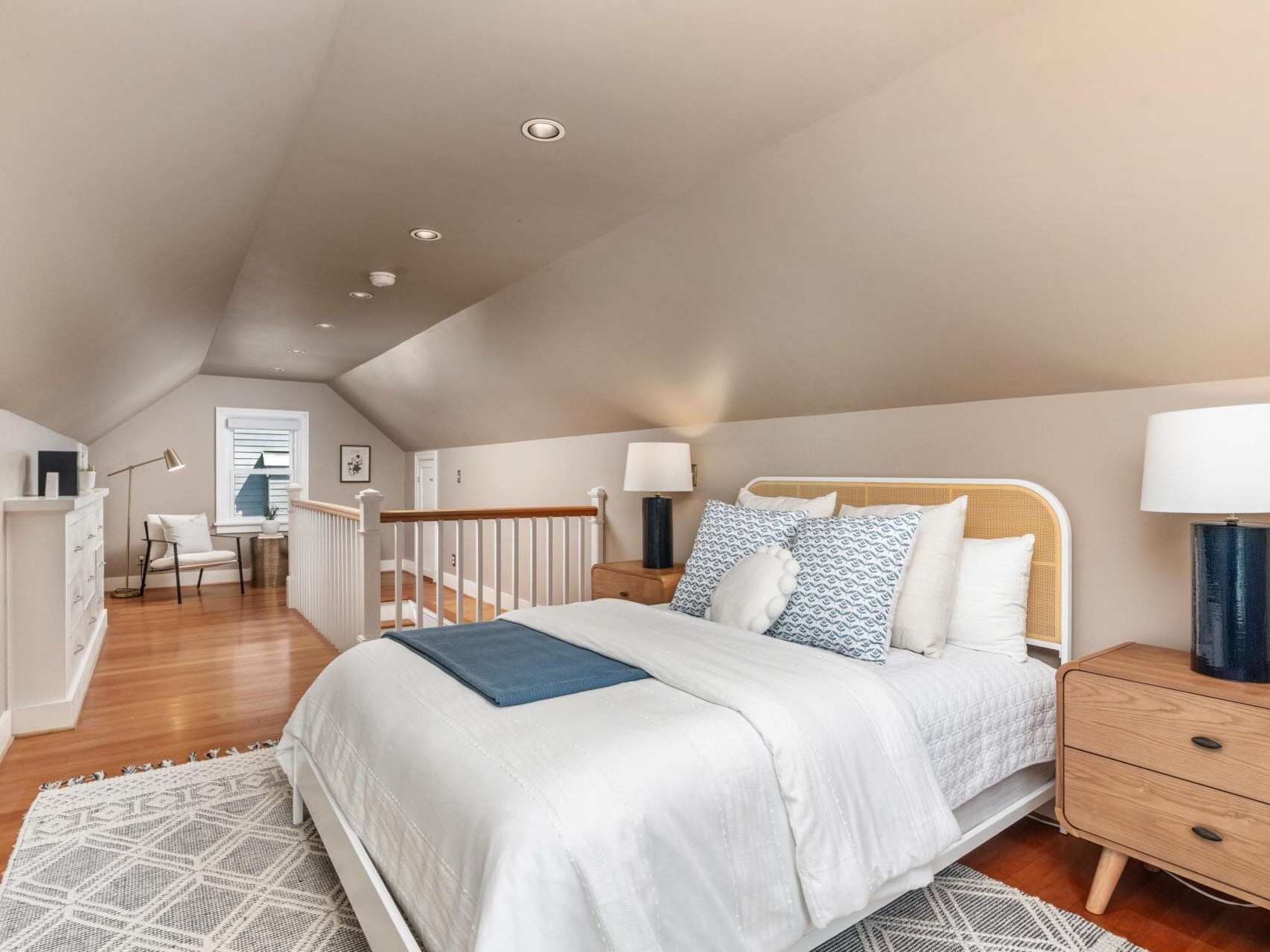 Bedroom in a finished attic with sloped ceiling, featuring a neatly made bed with white and blue bedding, wooden nightstands with lamps, a rug, dresser, and a small workspace by a window in the background.
