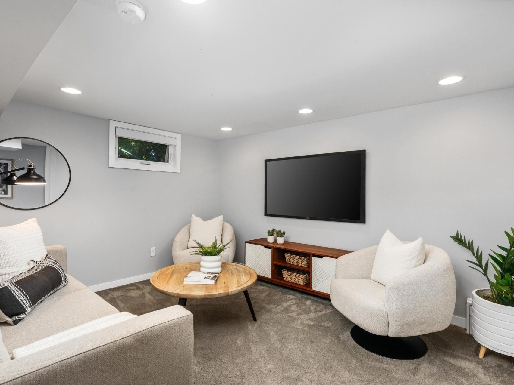 Modern, cozy living room with a beige sofa, two white armchairs, a round wooden coffee table, a TV mounted on the wall, a wooden media console, plants, and a round mirror, all set on a gray carpet.