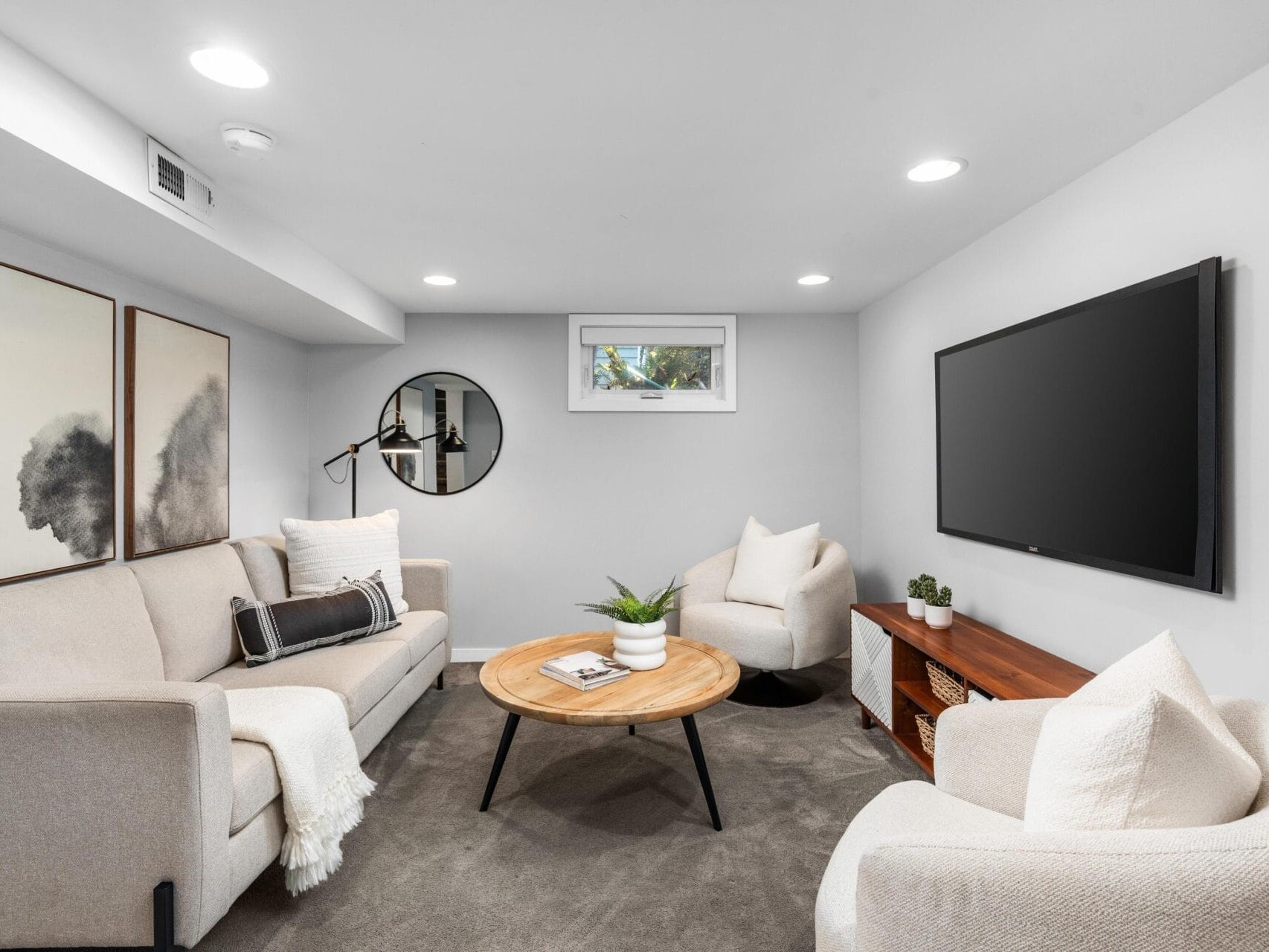 A modern living room with a light gray sofa, two white armchairs, a round wooden coffee table, wall art, a large TV mounted on the wall, and a small window letting in natural light.