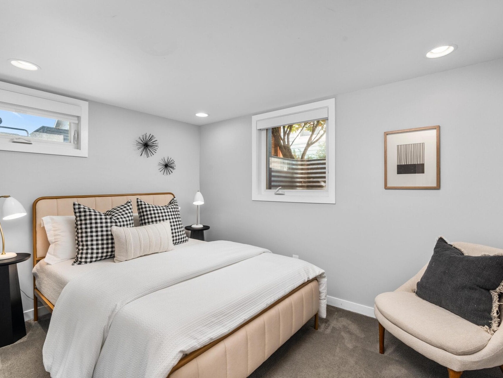 A modern bedroom with a beige bed, white bedding, black-and-white accent pillows, two black side tables with white lamps, a beige armchair with a dark pillow, and minimalist wall art under soft recessed lighting.