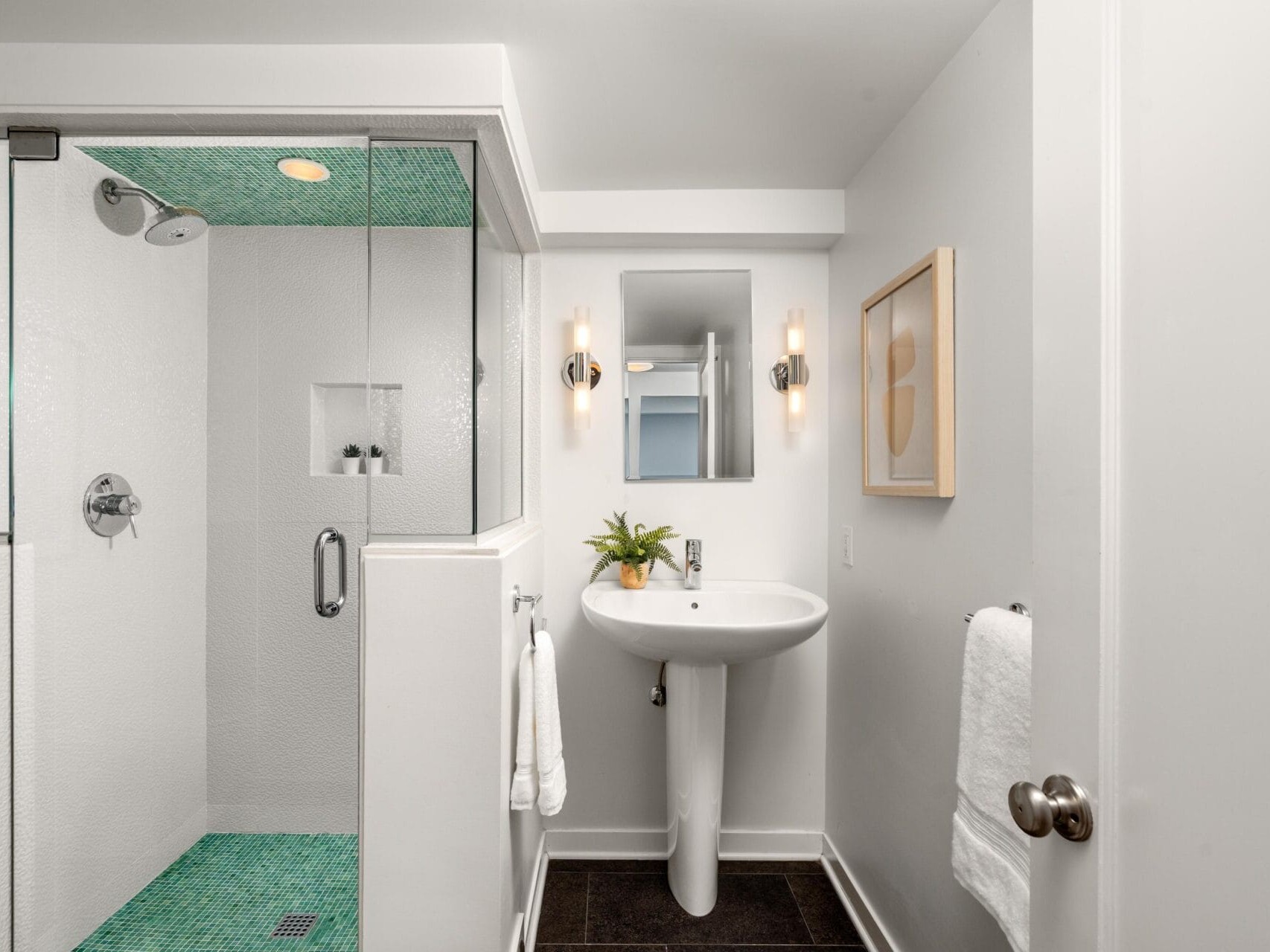 Modern bathroom with a glass-enclosed shower featuring teal tile floor, a pedestal sink, wall-mounted lights, a small mirror, a towel on a ring, and minimalist artwork on a white wall.