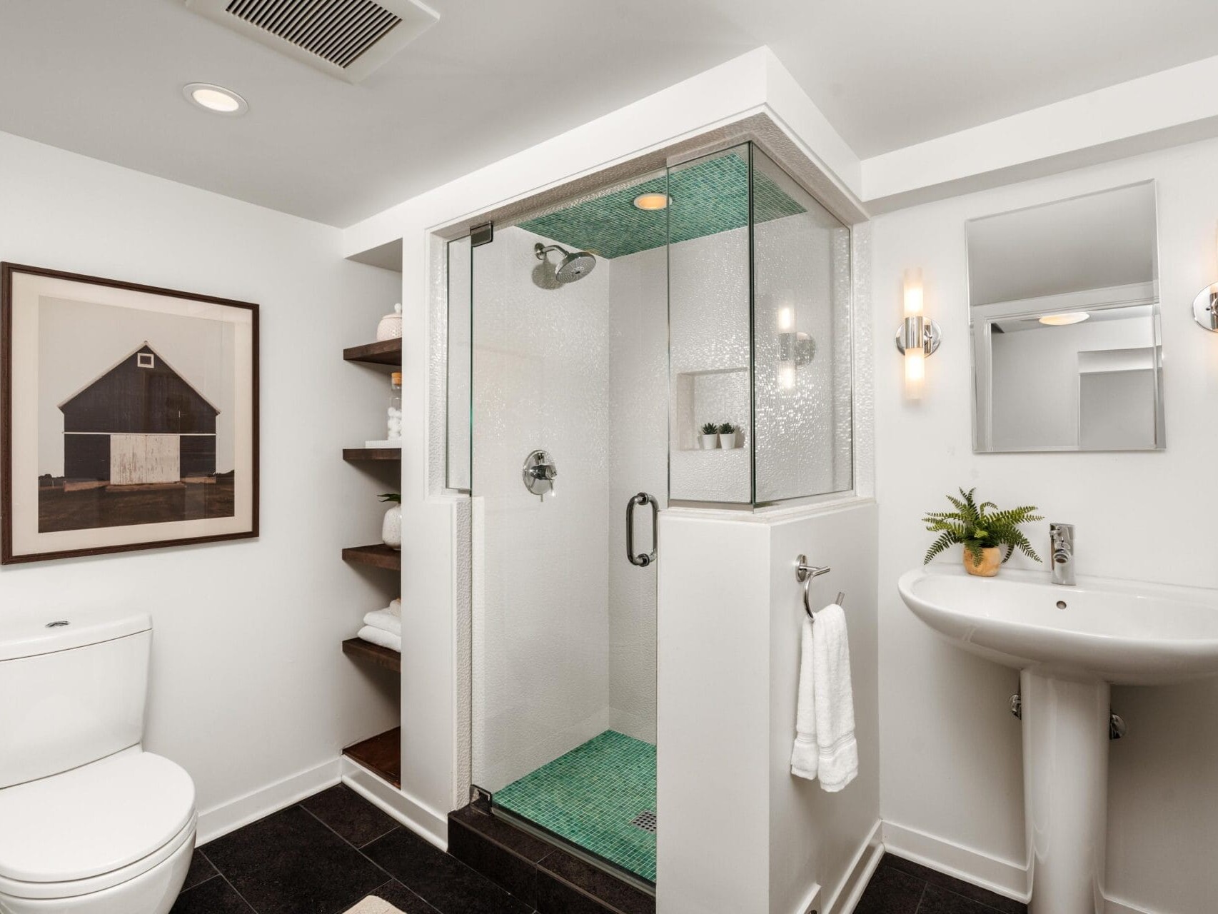 Modern bathroom with a glass shower, white pedestal sink, toilet, wall shelves with towels, a framed barn photo, and a small plant. The decor is minimal with light walls and dark floor tiles.