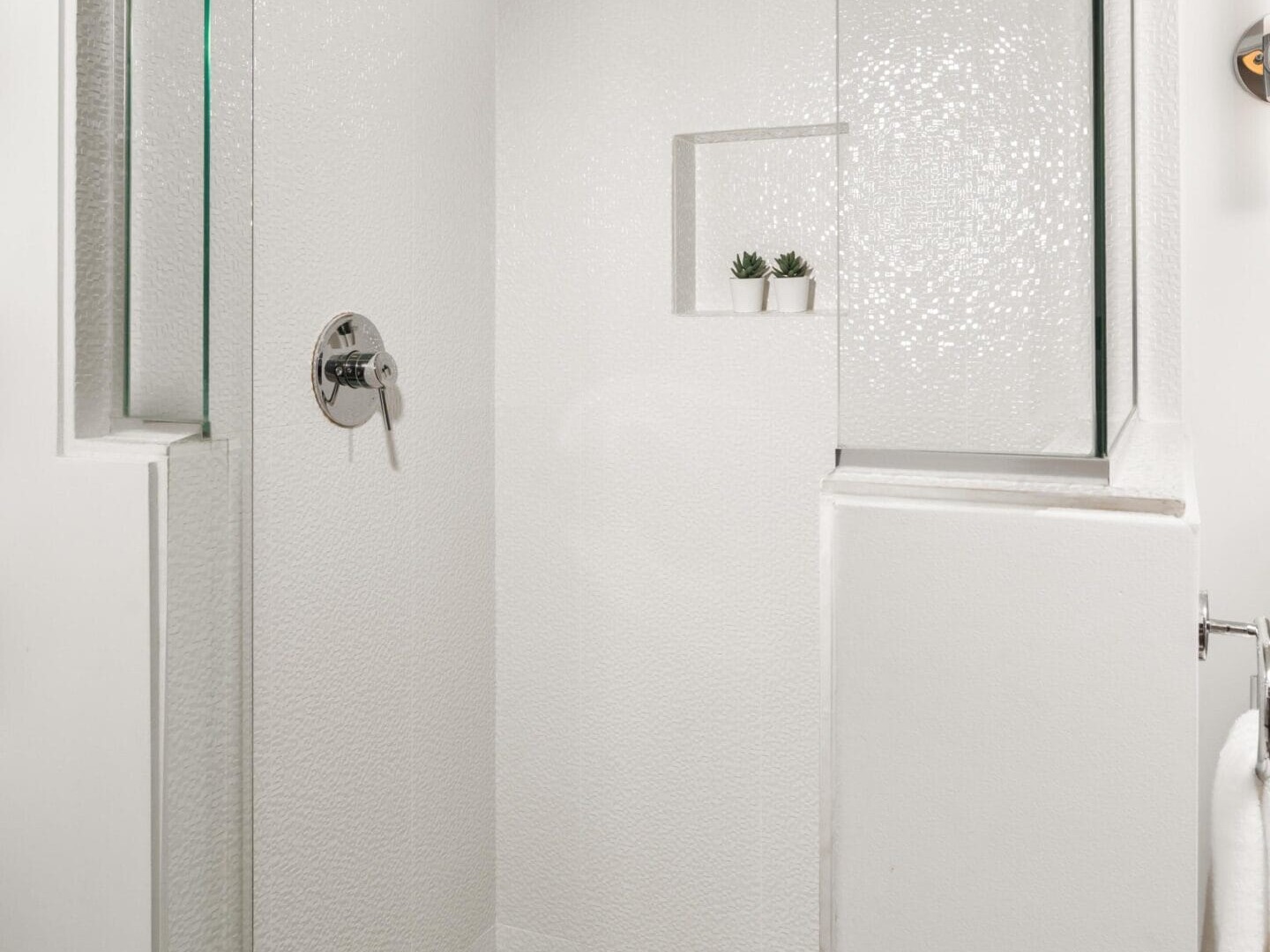 A modern glass shower with white textured walls and green mosaic tile floor and ceiling, featuring a built-in shelf holding two small potted plants. A silver showerhead and control handle are mounted on the wall.