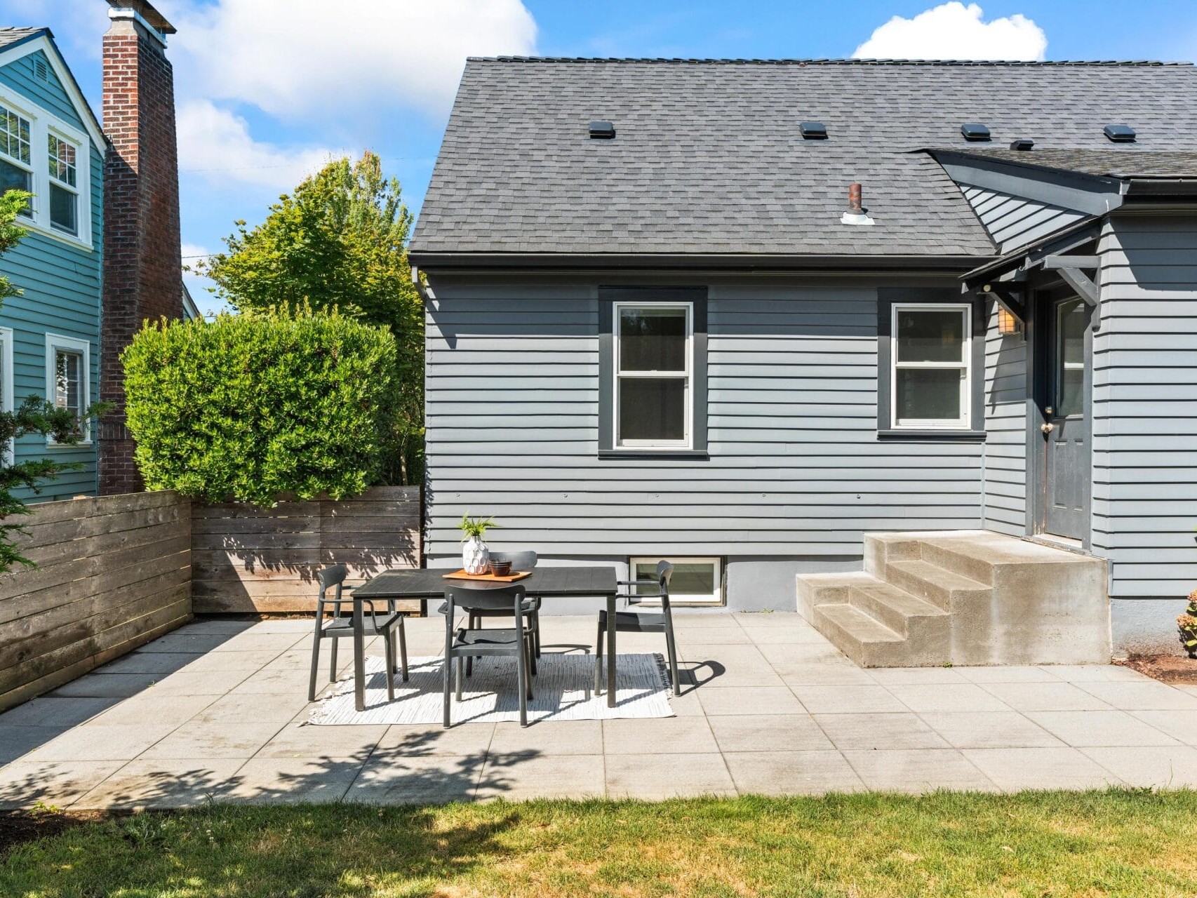 A small outdoor patio with a gray table and four chairs sits on a tiled area beside a gray house. Concrete steps lead to the door, and a wooden fence and green bushes border the space.