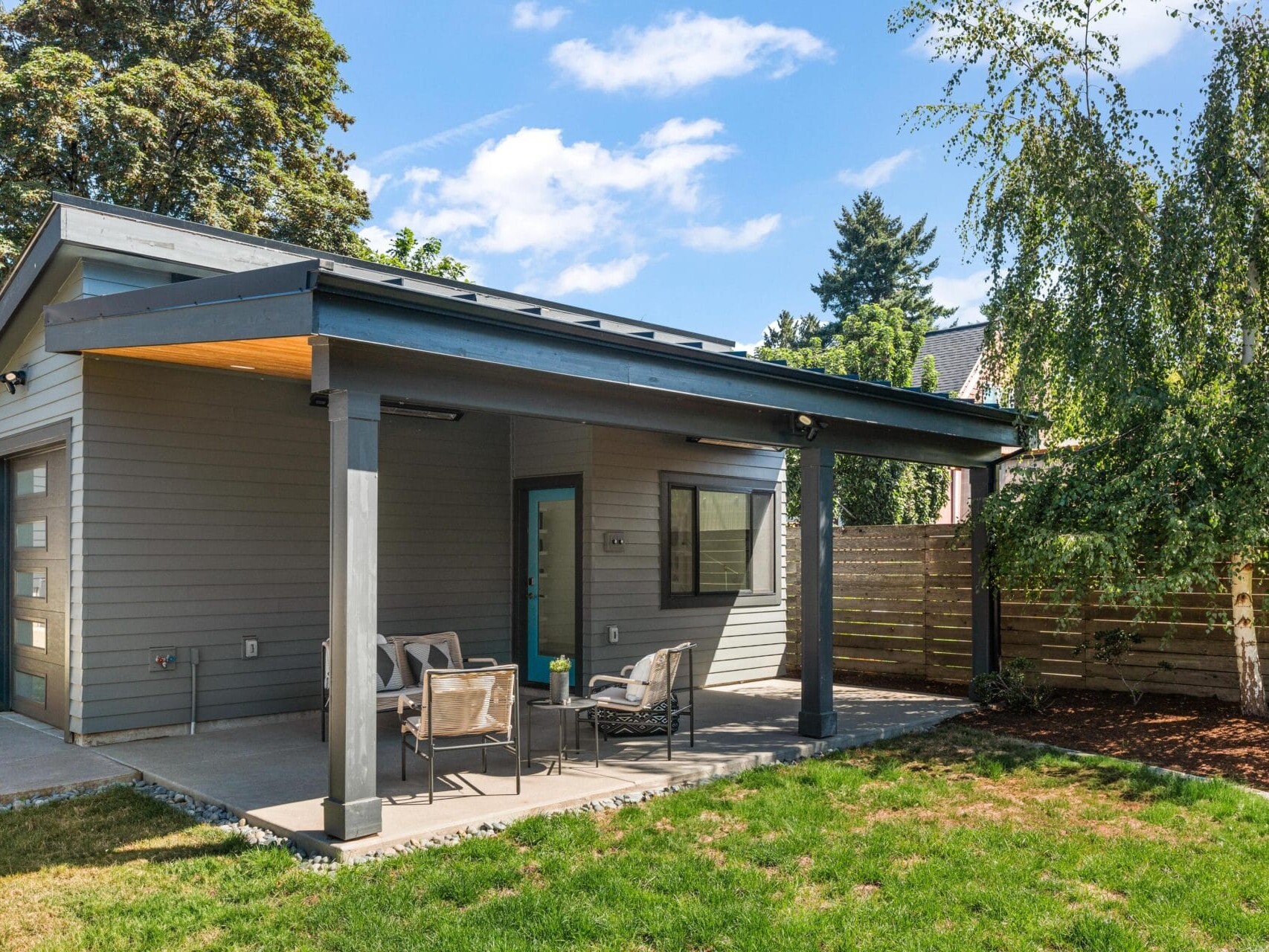 A modern house with gray siding and a sloped roof features a covered backyard patio with outdoor seating. The yard has green grass, a wooden fence, and trees under a blue sky with clouds.