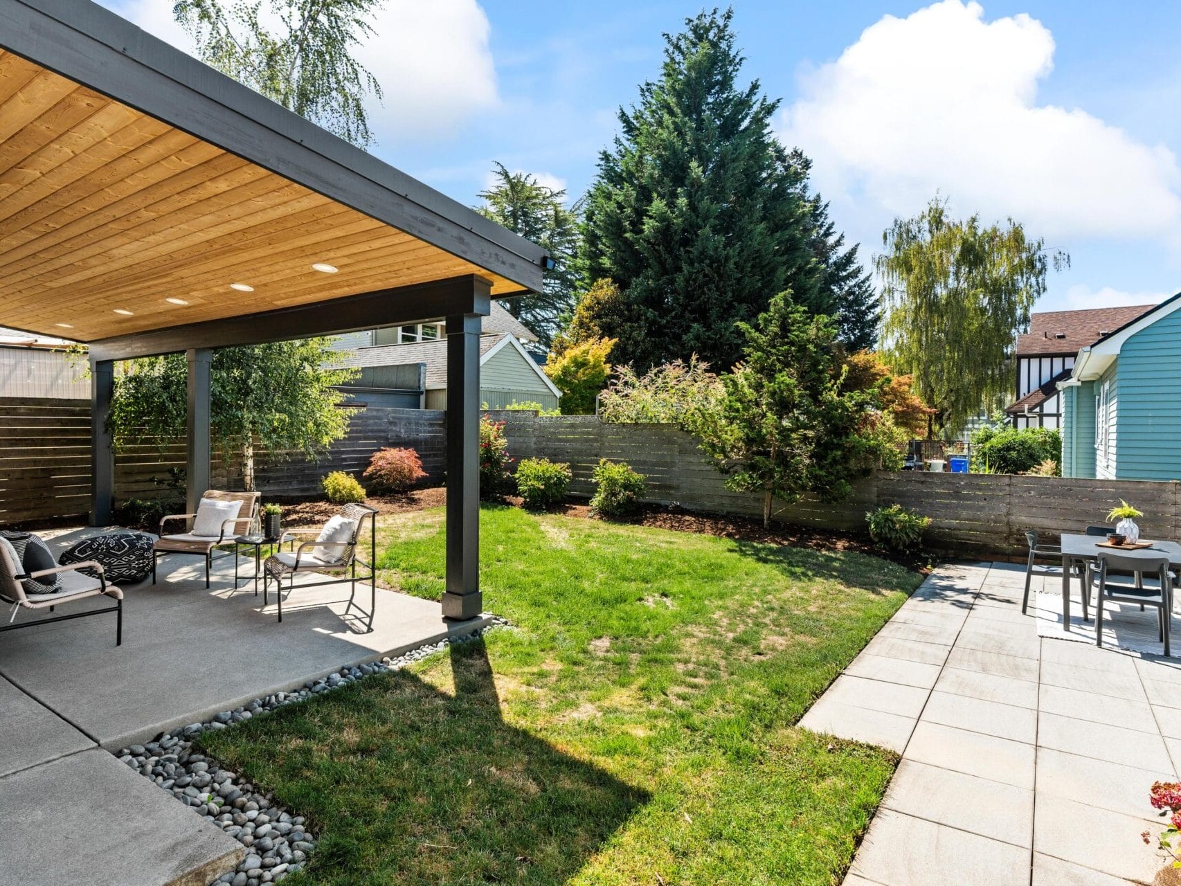 A backyard with a covered patio featuring outdoor seating, a grassy lawn, landscaped shrubs, a wood fence, and a tiled dining area with a table and chairs. Sunny weather with blue skies and light clouds.