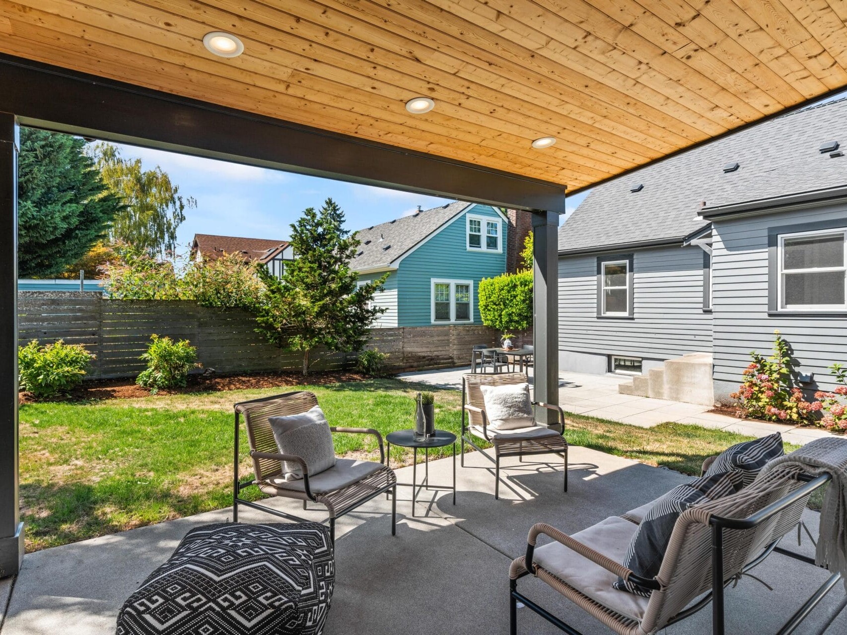 Covered patio with outdoor seating, poufs, and a small table, overlooking a grassy backyard with bushes and trees. Neighboring houses and a wooden fence are visible in the background under a blue sky.