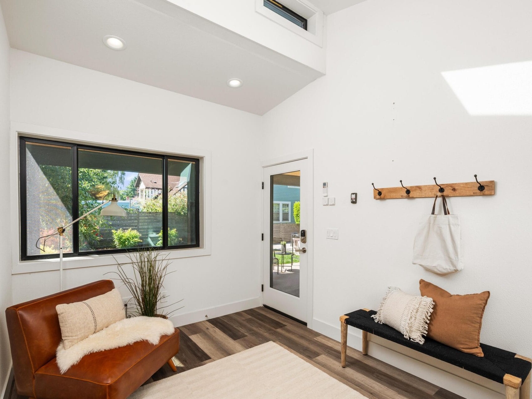 Bright entryway with a leather chair and a bench, both with neutral pillows. A wooden coat rack with a tote bag hangs on the white wall. Large window and glass door reveal a sunny outdoor scene.