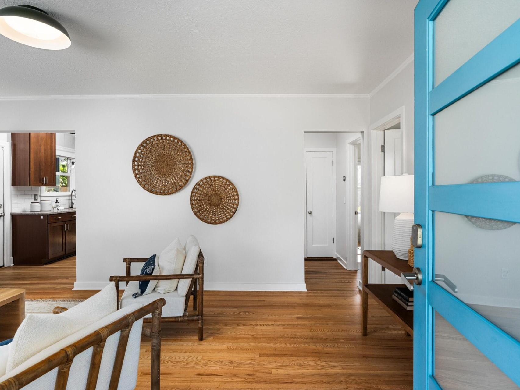 Bright living room with wooden floors, white walls, and modern furnishings. A blue door is open, revealing chairs, a console table with a lamp, and two woven wall hangings; a kitchen is visible in the background.