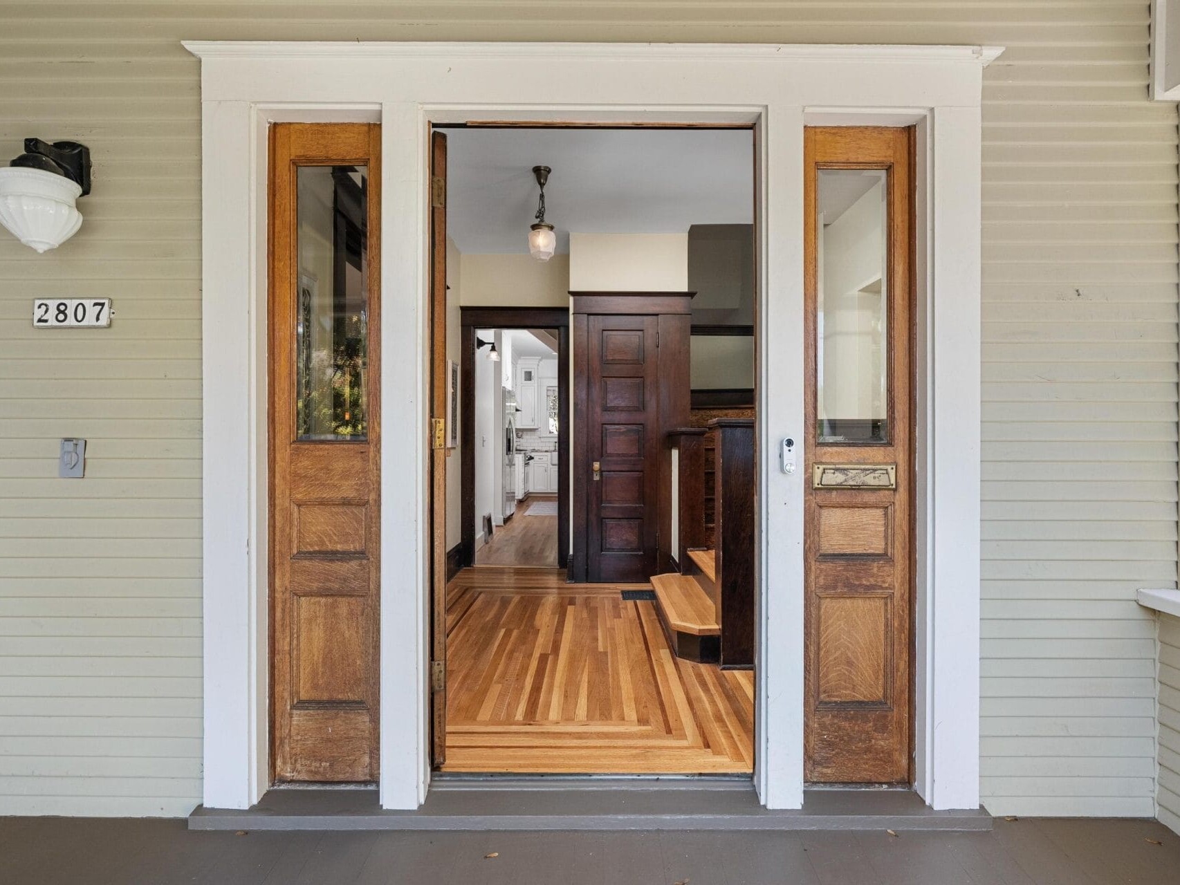 A front porch view of a house with double wooden doors open, revealing a hallway with wood floors and a dark wooden door inside. The exterior has light-colored siding and a house number “2807” is visible on the left.