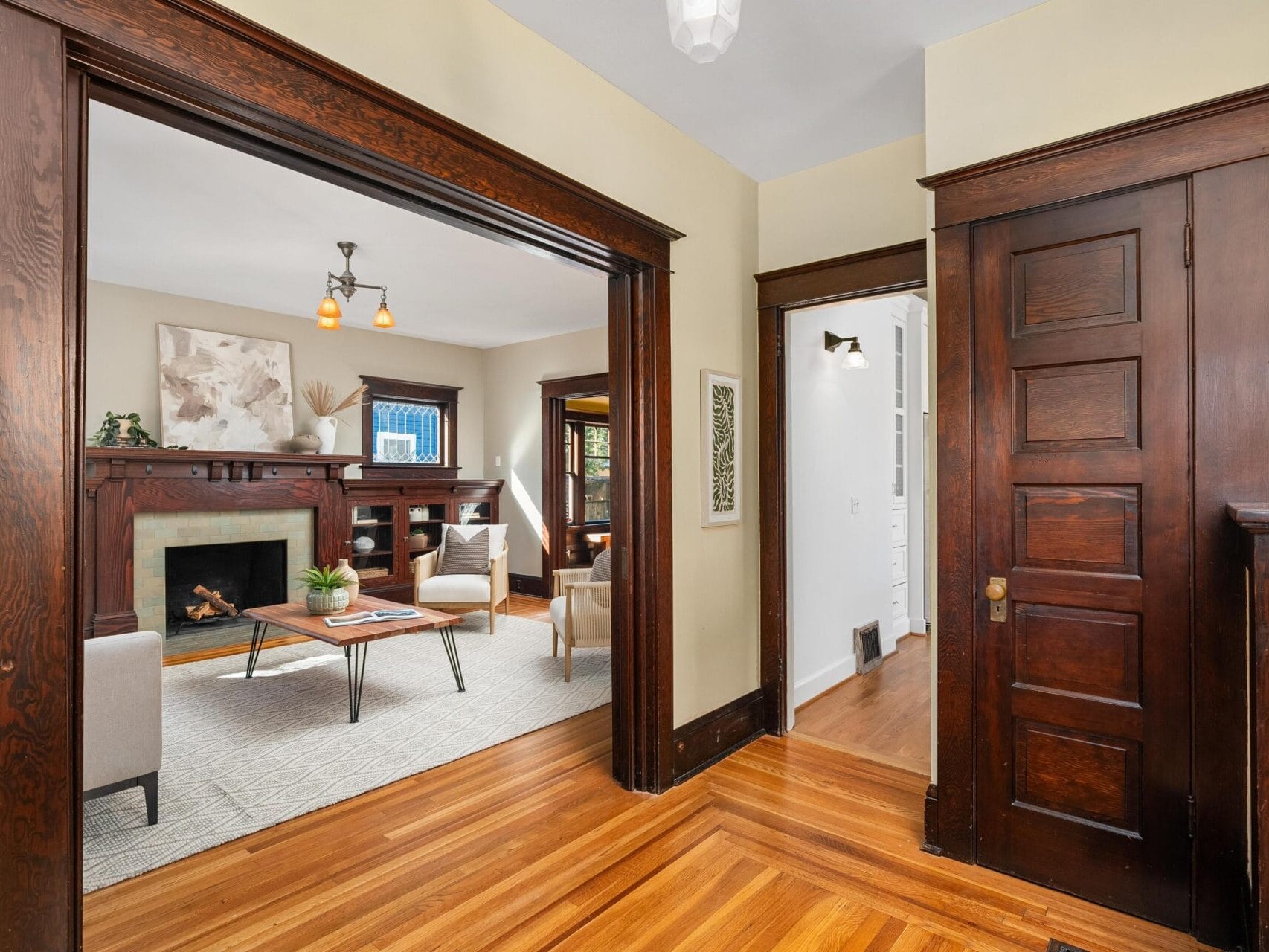 Interior view of a home with hardwood floors, dark wood trim, and a living room featuring a fireplace, light-colored armchairs, a coffee table, and large windows letting in natural light.