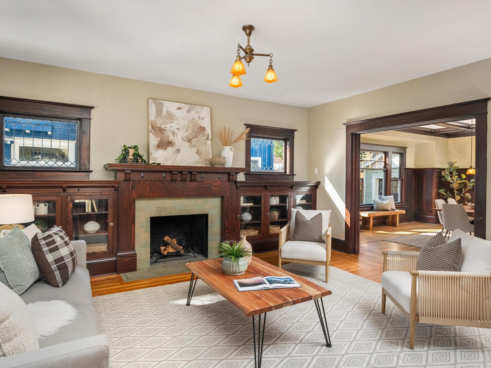 A cozy living room with a lit fireplace, wooden built-in shelves, beige walls, gray sofa, armchair, coffee table with a book and plant, and a doorway leading to a sunlit adjacent room.