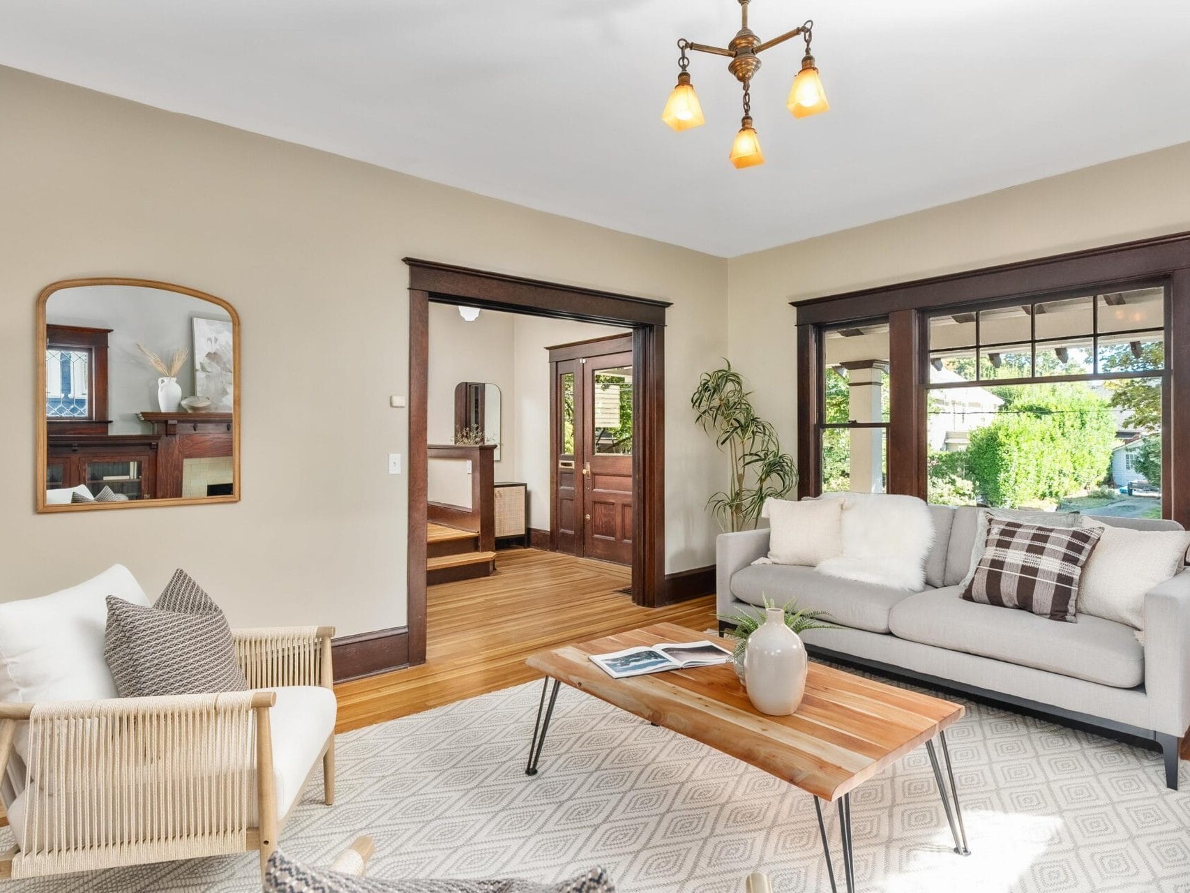 A bright living room with a light sofa, armchair, wooden coffee table, patterned rug, and large windows. The walls are beige, and dark wood trim frames the windows and doorway. A mirror hangs on the wall.