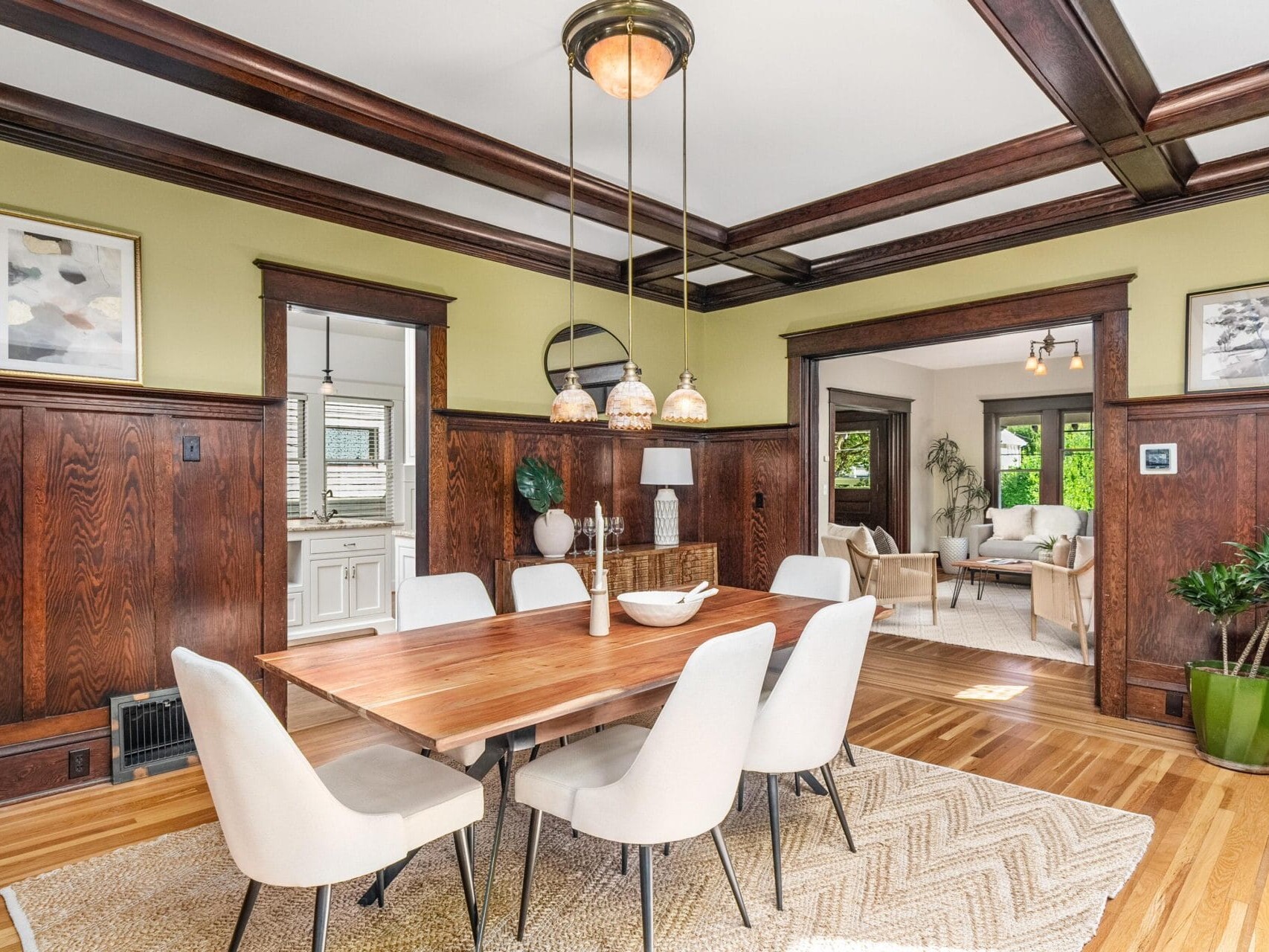 A dining room with a wooden table, six white chairs, hardwood floors, and dark wood paneling. Light fixtures hang above the table, and a living room with natural light is visible through a wide doorway.