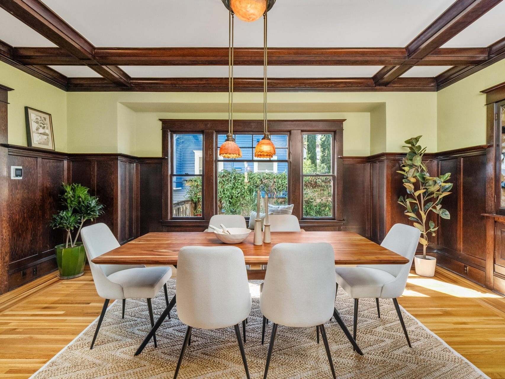 A dining room with a wooden table, six white chairs, pendant lights above, large windows, wood paneling on the walls, hardwood floors, potted plants, and a beige patterned rug.