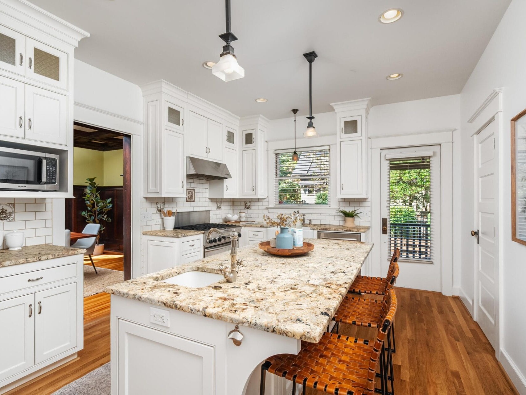 Bright, modern kitchen with white cabinets, granite countertops, an island with wicker barstools, stainless steel appliances, and pendant lighting. Large windows let in natural light, and wood flooring adds warmth.