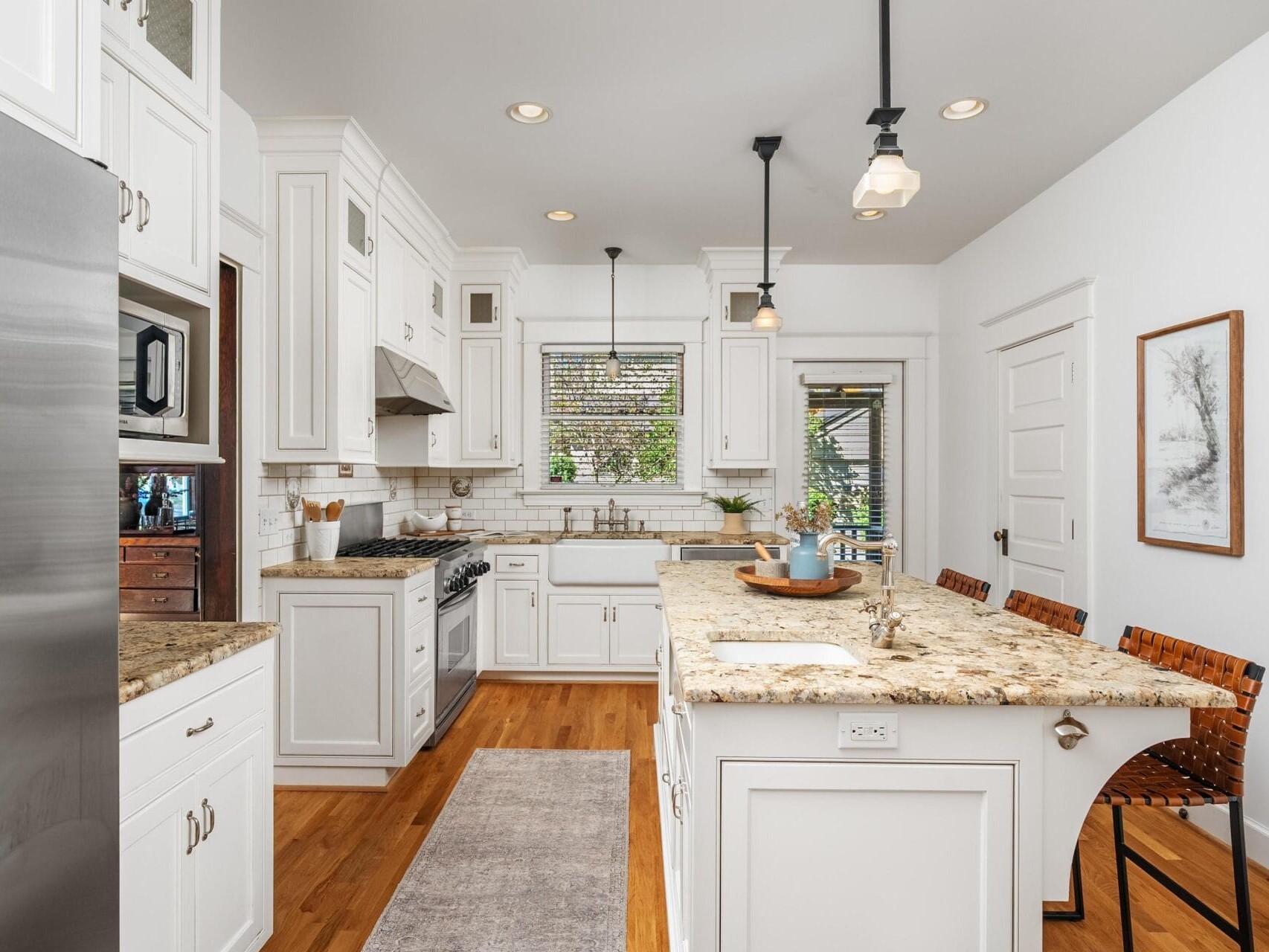 Bright, modern kitchen with white cabinets, granite countertops, stainless steel appliances, a center island with woven bar stools, hardwood floors, pendant lights, and a window above the sink letting in natural light.