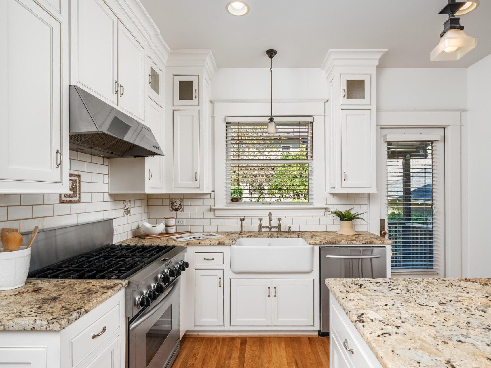 Bright kitchen with white cabinets, granite countertops, a farmhouse sink, stainless steel stove, dishwasher, tile backsplash, and wooden floors. A window above the sink lets in natural light.