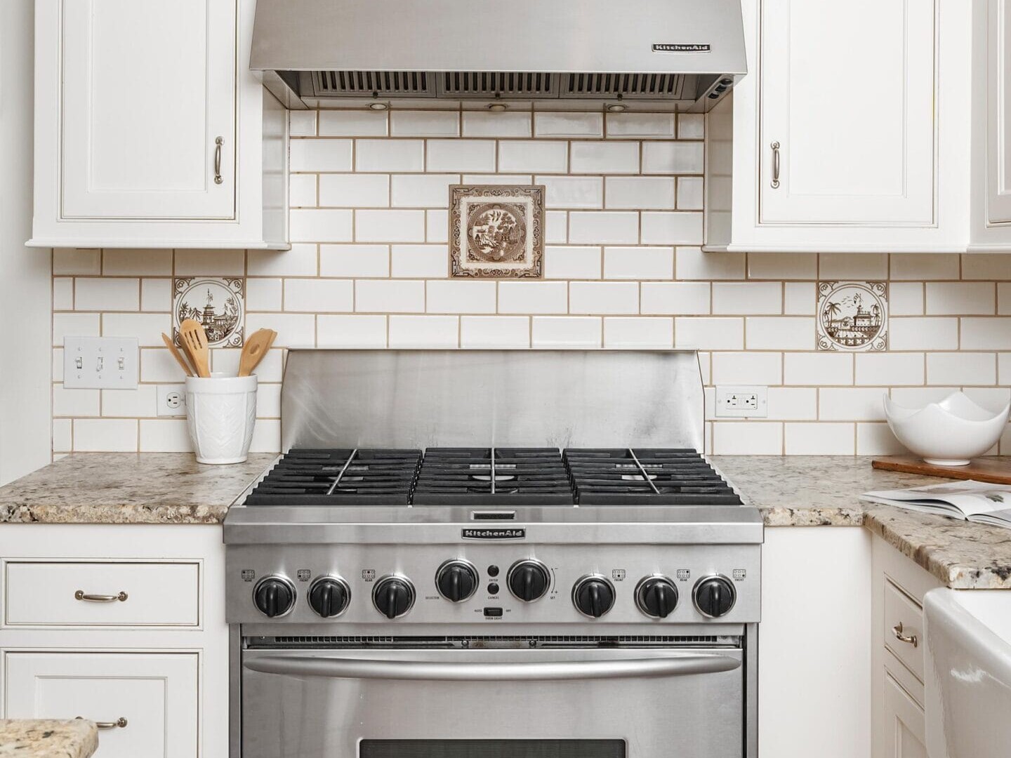 A modern kitchen with white cabinets, subway tile backsplash, a stainless steel gas stove and oven, a metal range hood, and granite countertops with utensils and a bowl on the sides.