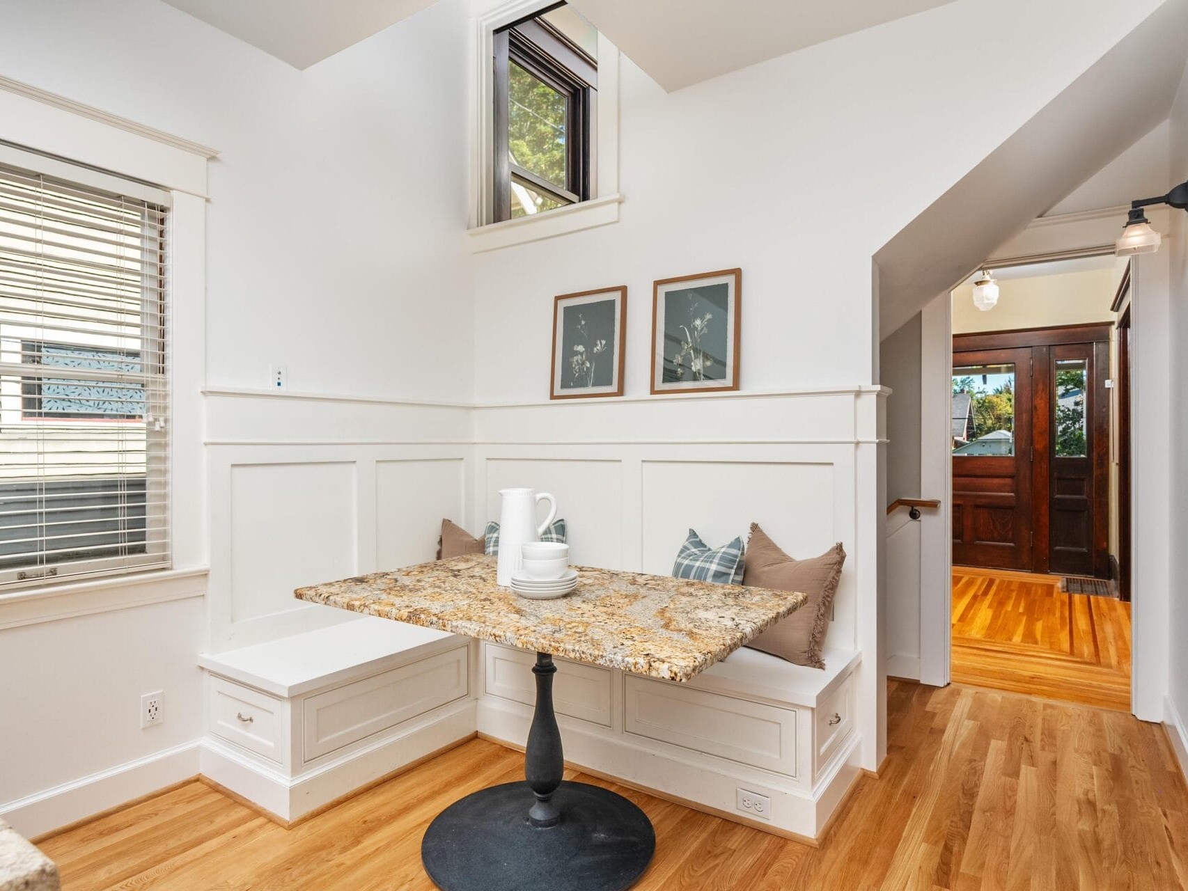 A cozy breakfast nook with a granite table, built-in white bench seating, neutral cushions, and wall art. Sunlight streams through a window, and a hallway leads to a wooden front door.