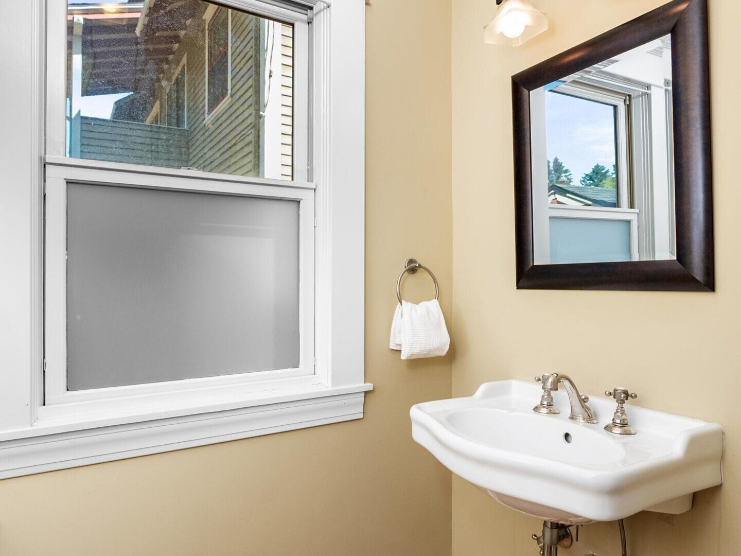 A small bathroom corner with a white wall-mounted sink, a rectangular mirror with a dark frame, a two-light fixture above, a towel ring with a white towel, yellow walls, wood flooring, and a window with frosted glass.