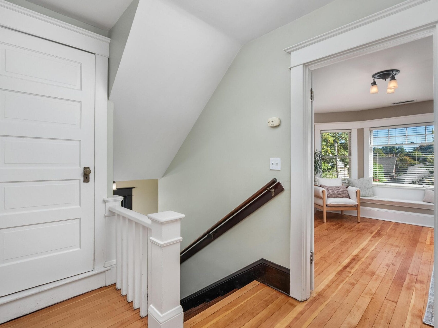 A hallway with light wood floors, a white door on the left, a staircase with a wooden handrail, and an open doorway leading to a bright room with large windows and a white armchair.