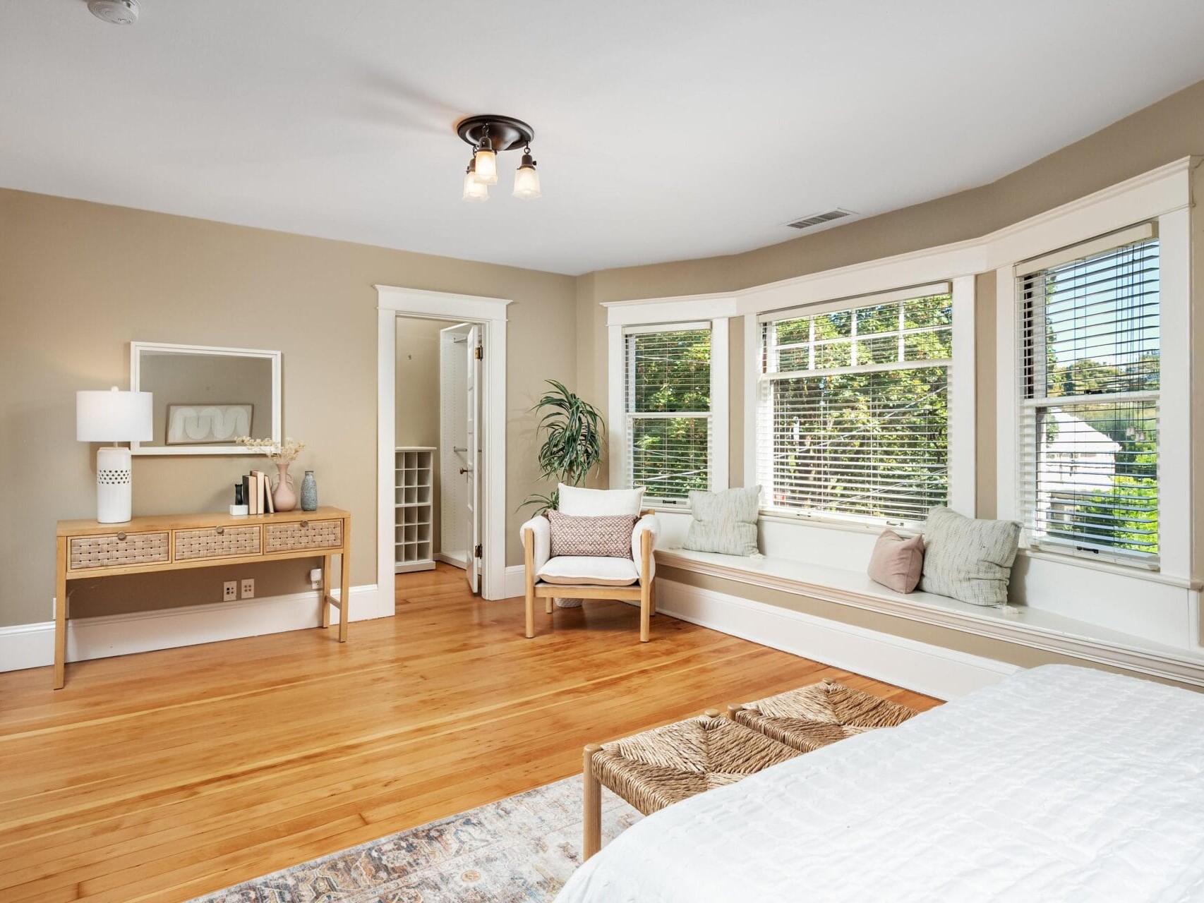 Bright bedroom with light wood floors, beige walls, a bed, a window bench with cushions, a chair, a writing desk with a mirror, and large windows letting in natural light with a view of trees outside.